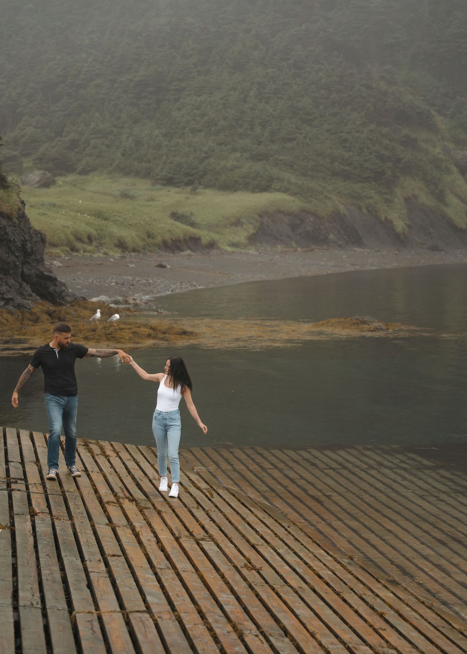 A couple holds hands while standing on a wooden dock overlooking a misty lake surrounded by green hills.