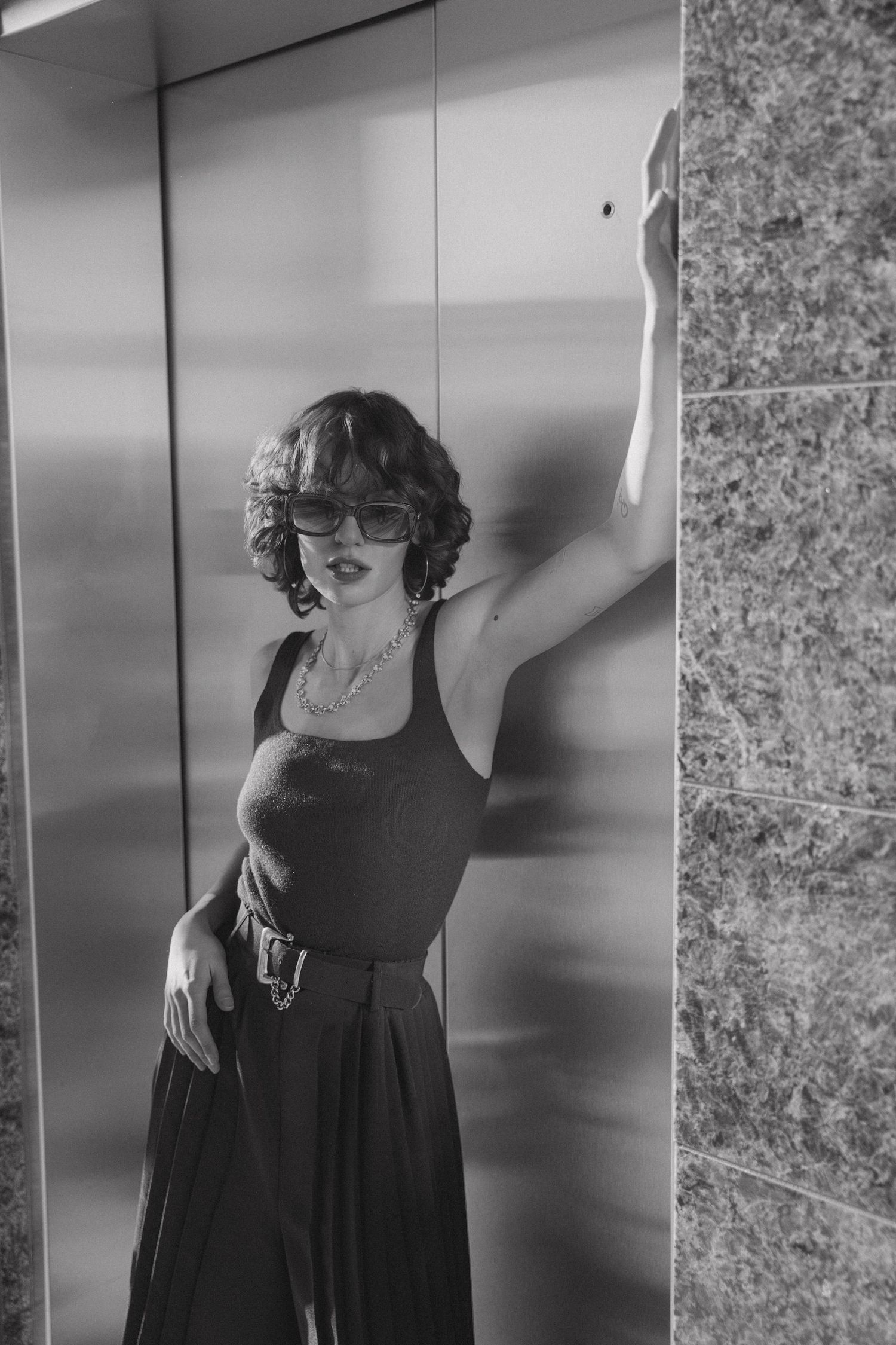 A black and white fashion portrait of someone in a dark dress and sunglasses posing against a light background.