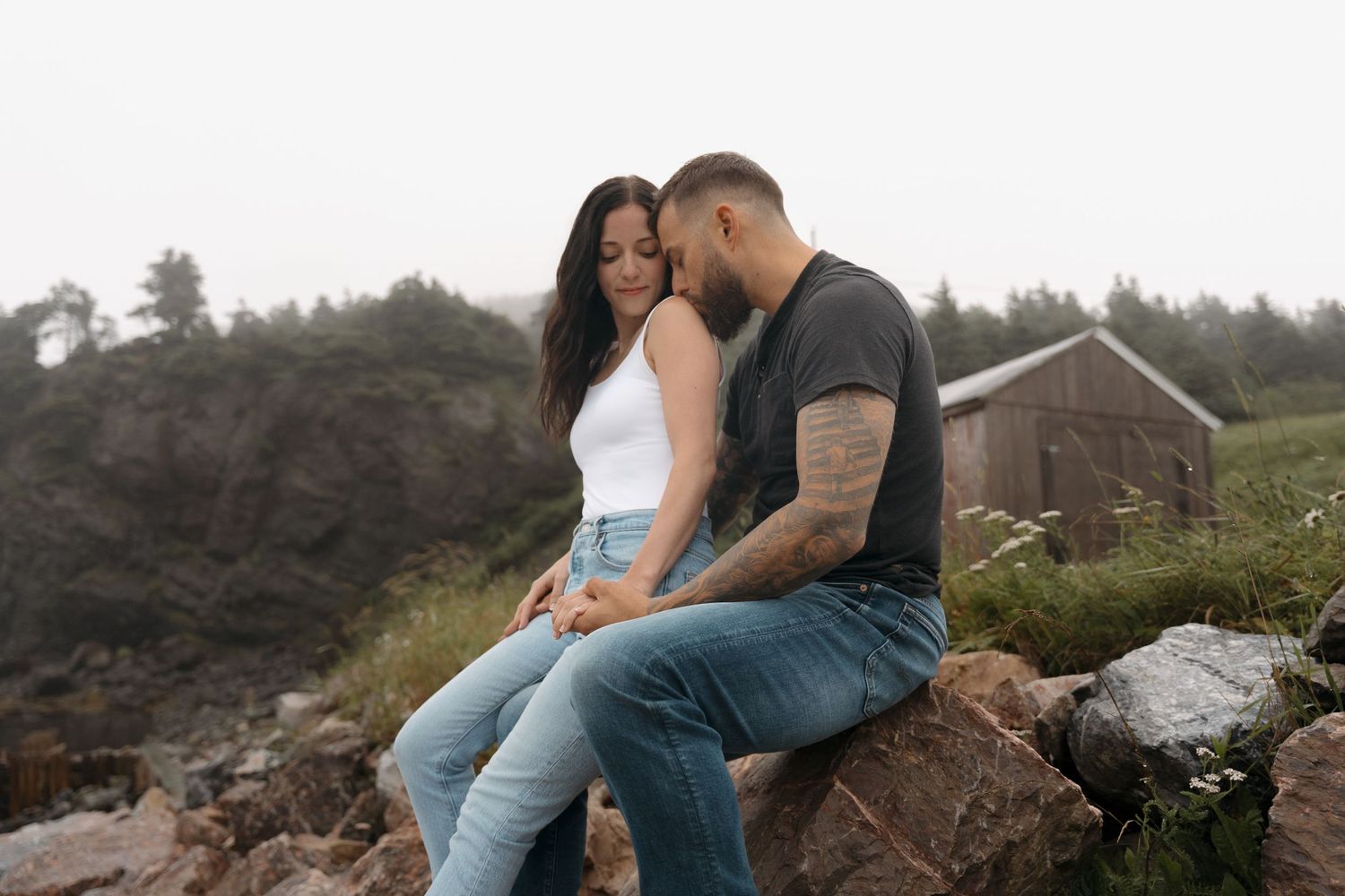 A romantic couple embraces on a rocky cliff overlooking a misty forest landscape during golden hour.