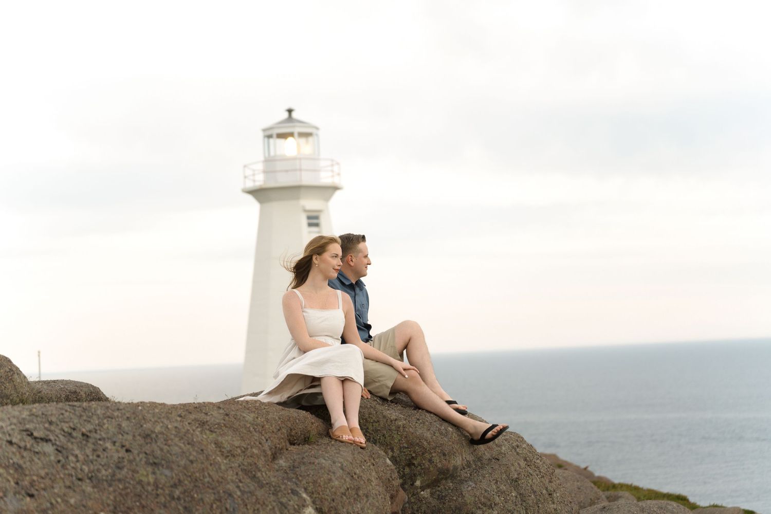 A couple sits on coastal rocks near a white lighthouse overlooking the ocean at sunset.