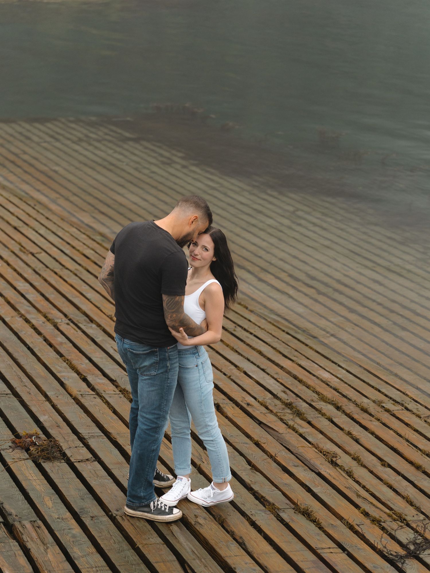 A couple shares an intimate moment standing together on a wooden dock overlooking water.
