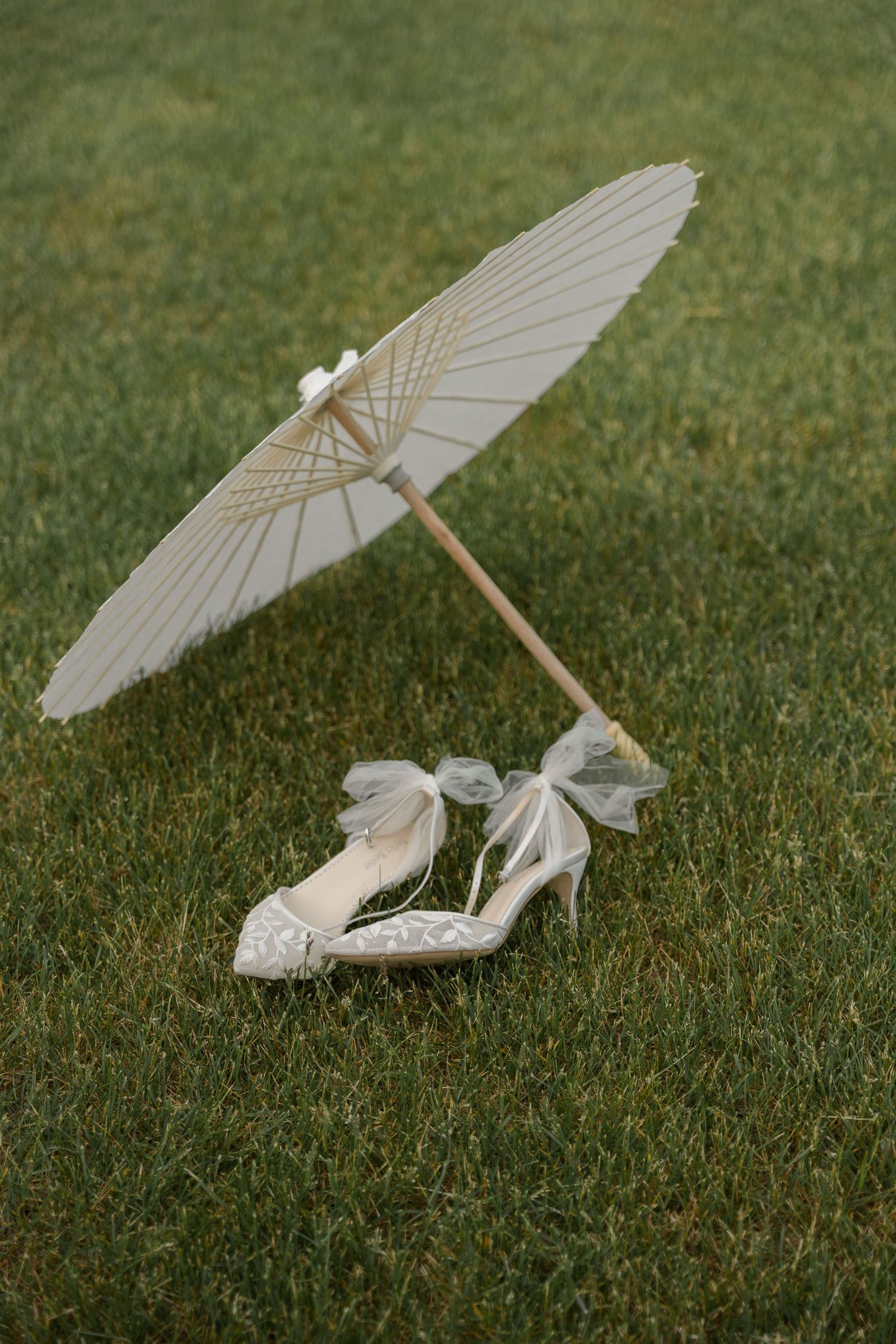 White parasol and lace wedding shoes arranged on grass for artistic photo.