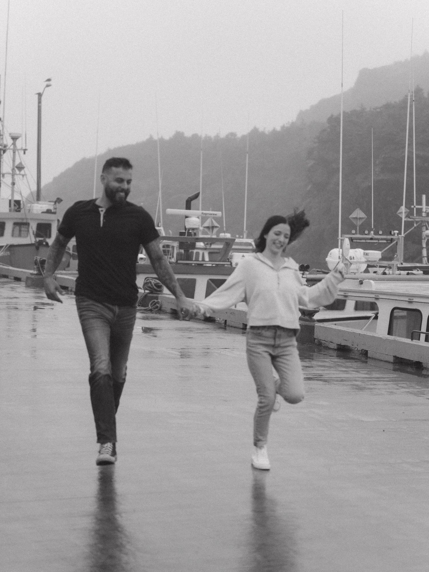 A couple runs and plays together on a wet dock with boats and mountains in the background, captured in black and white.