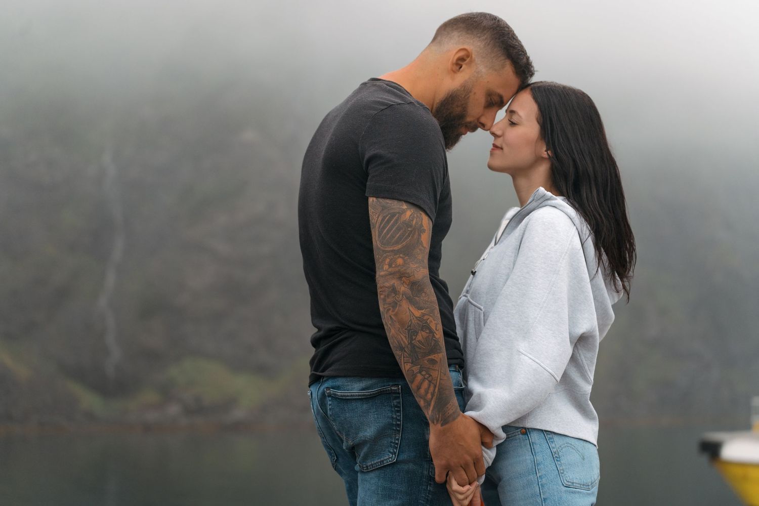 A romantic couple holds hands and shares an intimate moment against a misty mountain backdrop.