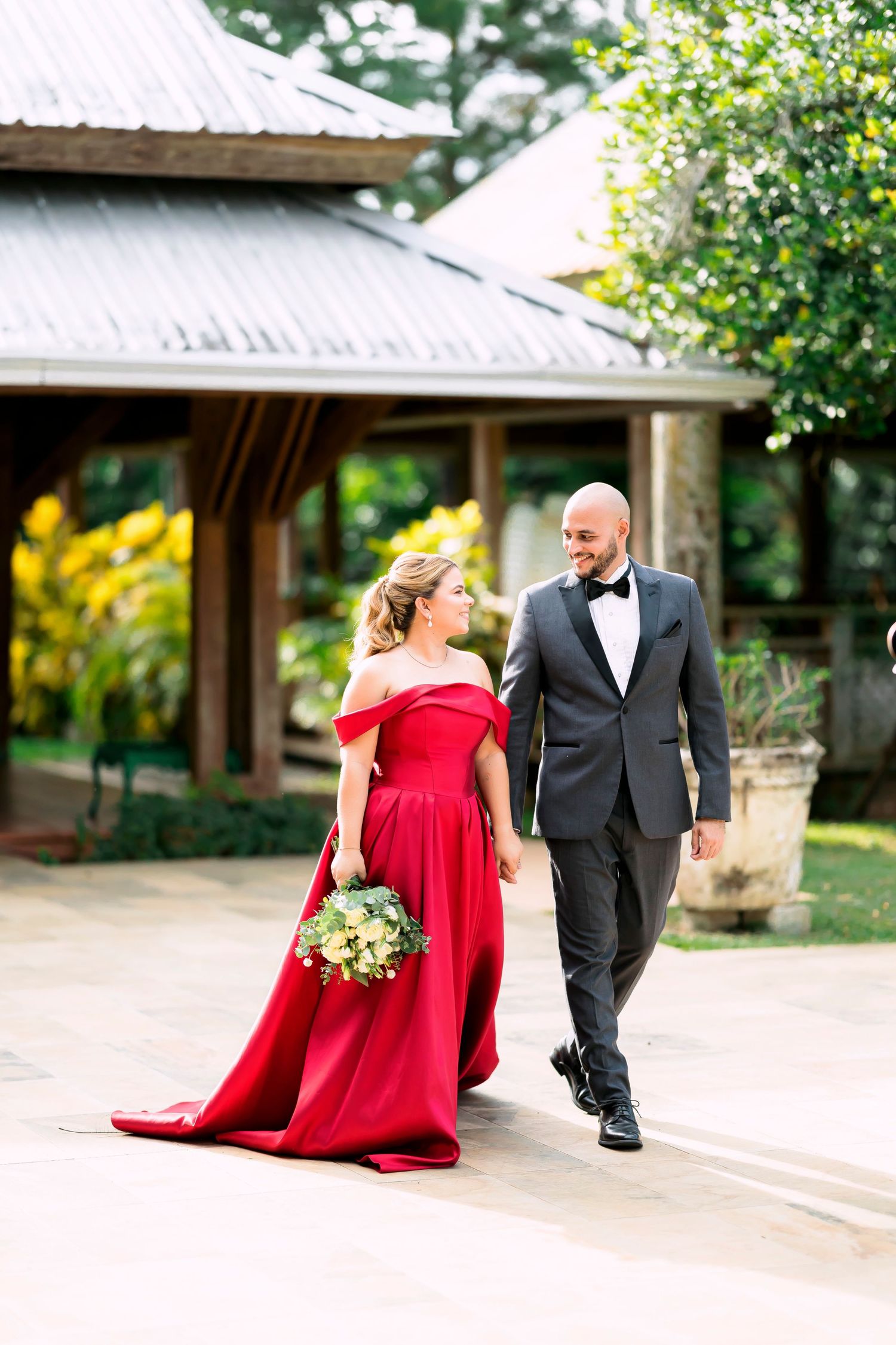 A couple walks together on garden path, with bride wearing striking red off-shoulder gown.