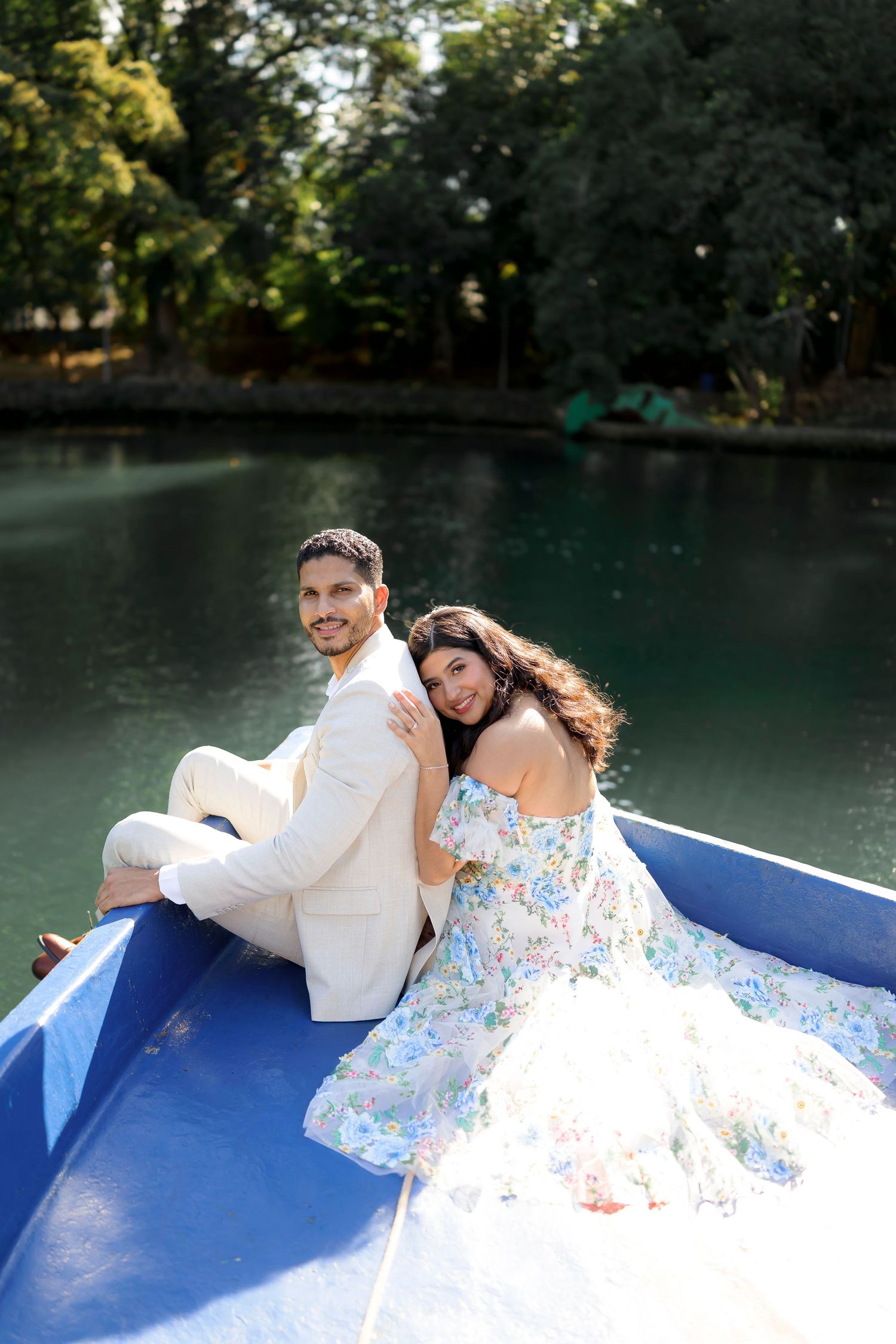 A couple in romantic attire sitting back to back on a blue boat in a serene lake surrounded by trees.