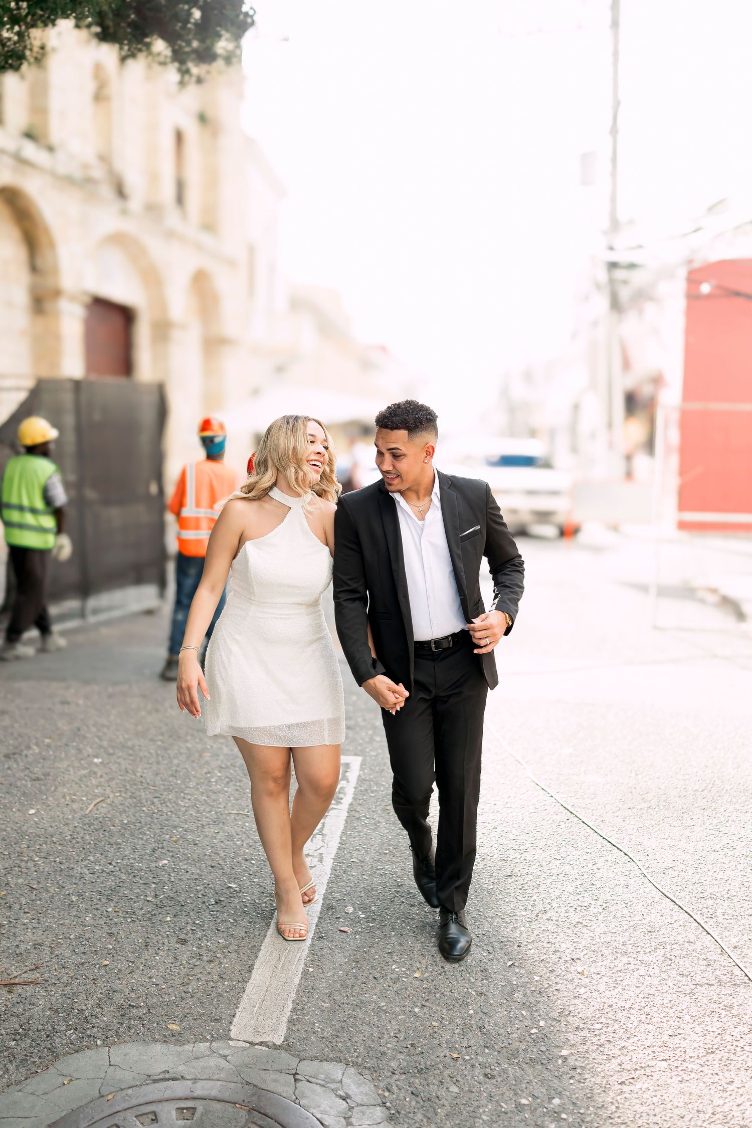 Couple walking together on urban street with construction workers in background.