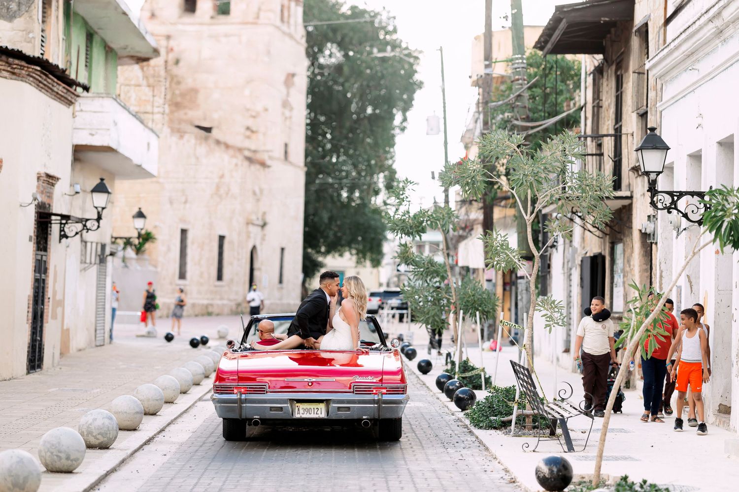 A vintage red convertible car drives through a charming European-style street with stone buildings and lamp posts.