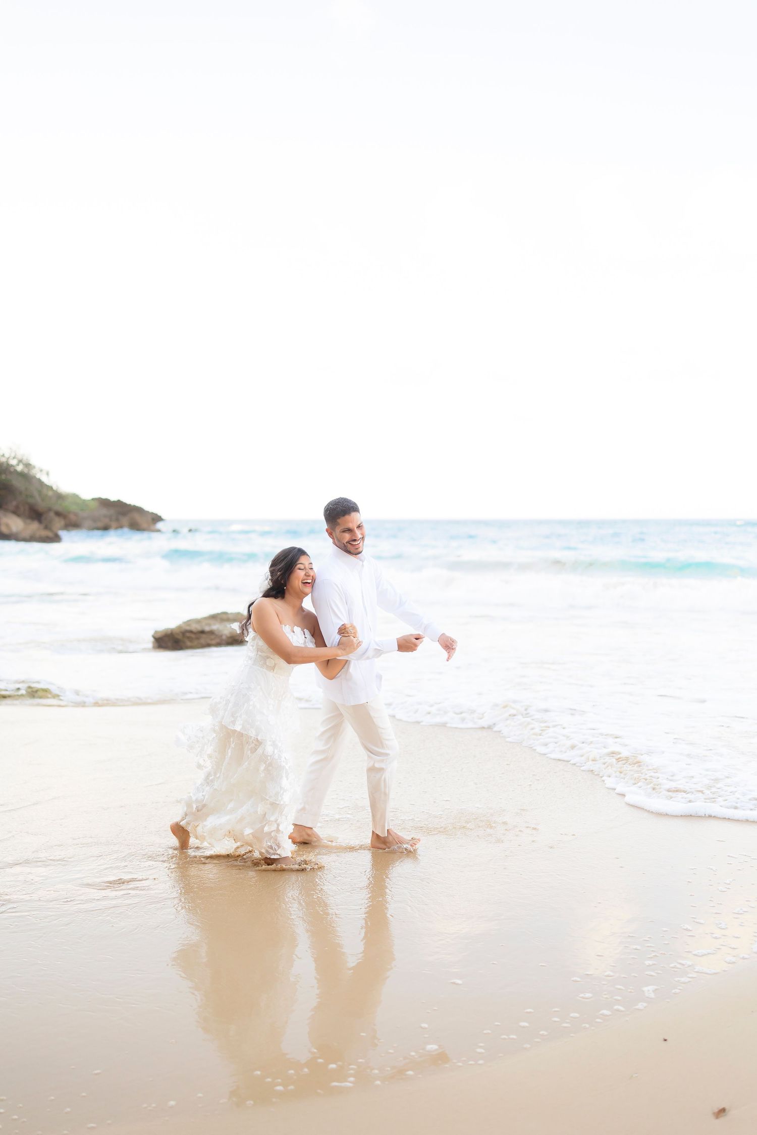 A joyful couple in white attire dances together on a scenic beach with waves and rocky coastline in the background.