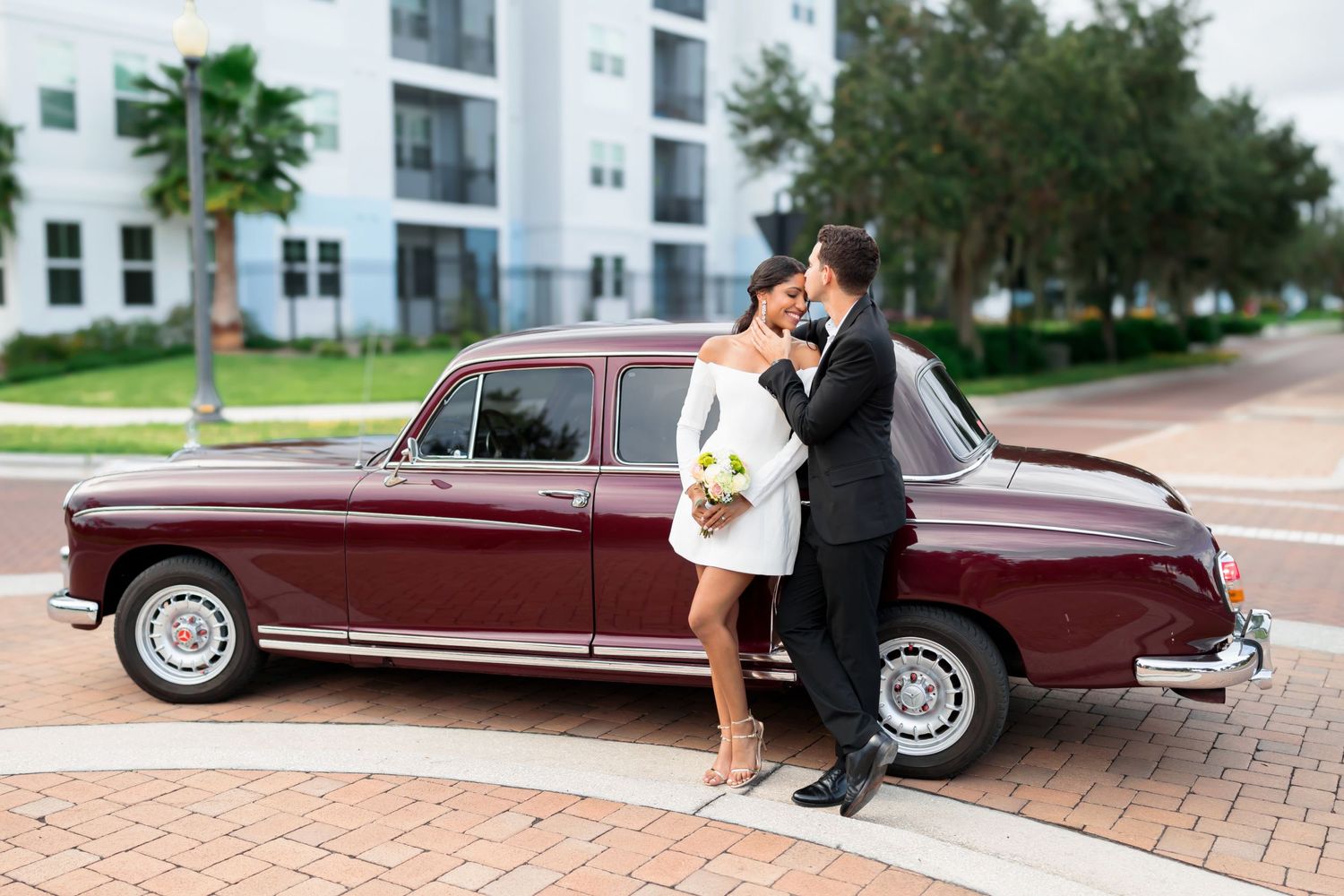 Couple poses by vintage burgundy car for wedding photos on brick pavement.