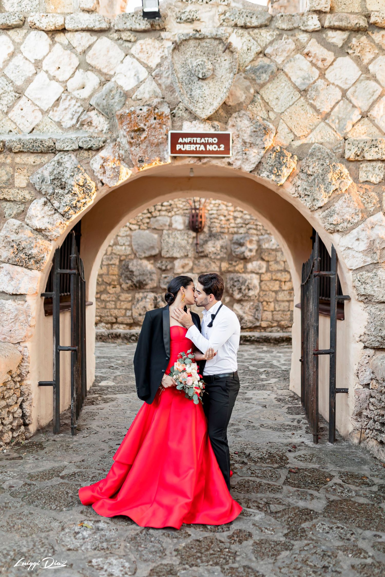 Couple in formal attire embracing under stone archway with red gown and bouquet.