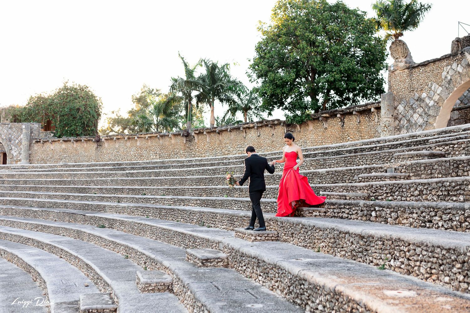 Couple walking up stone amphitheater steps with person in flowing red dress.