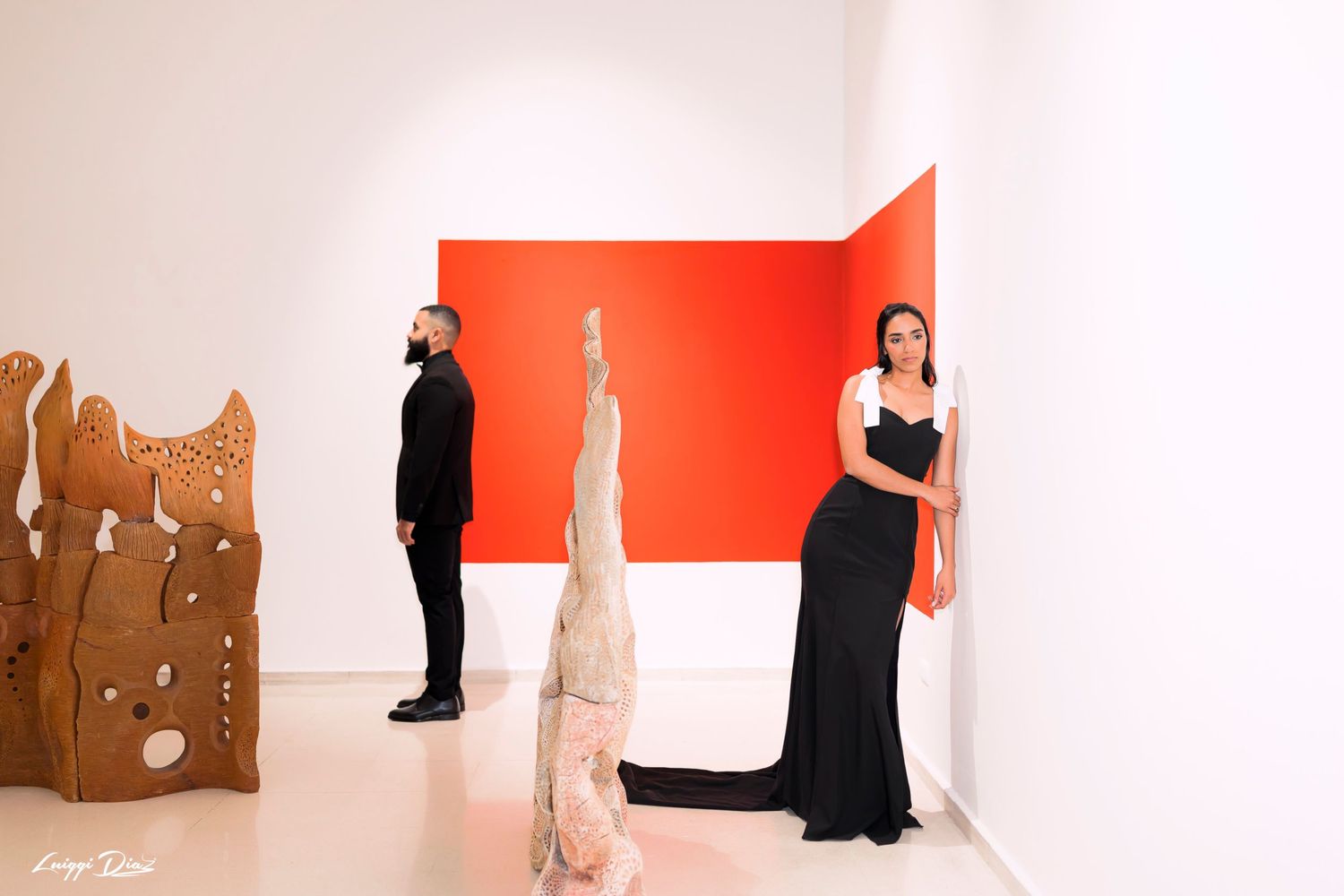Three figures in formal attire pose against a red and white wall in an art gallery.