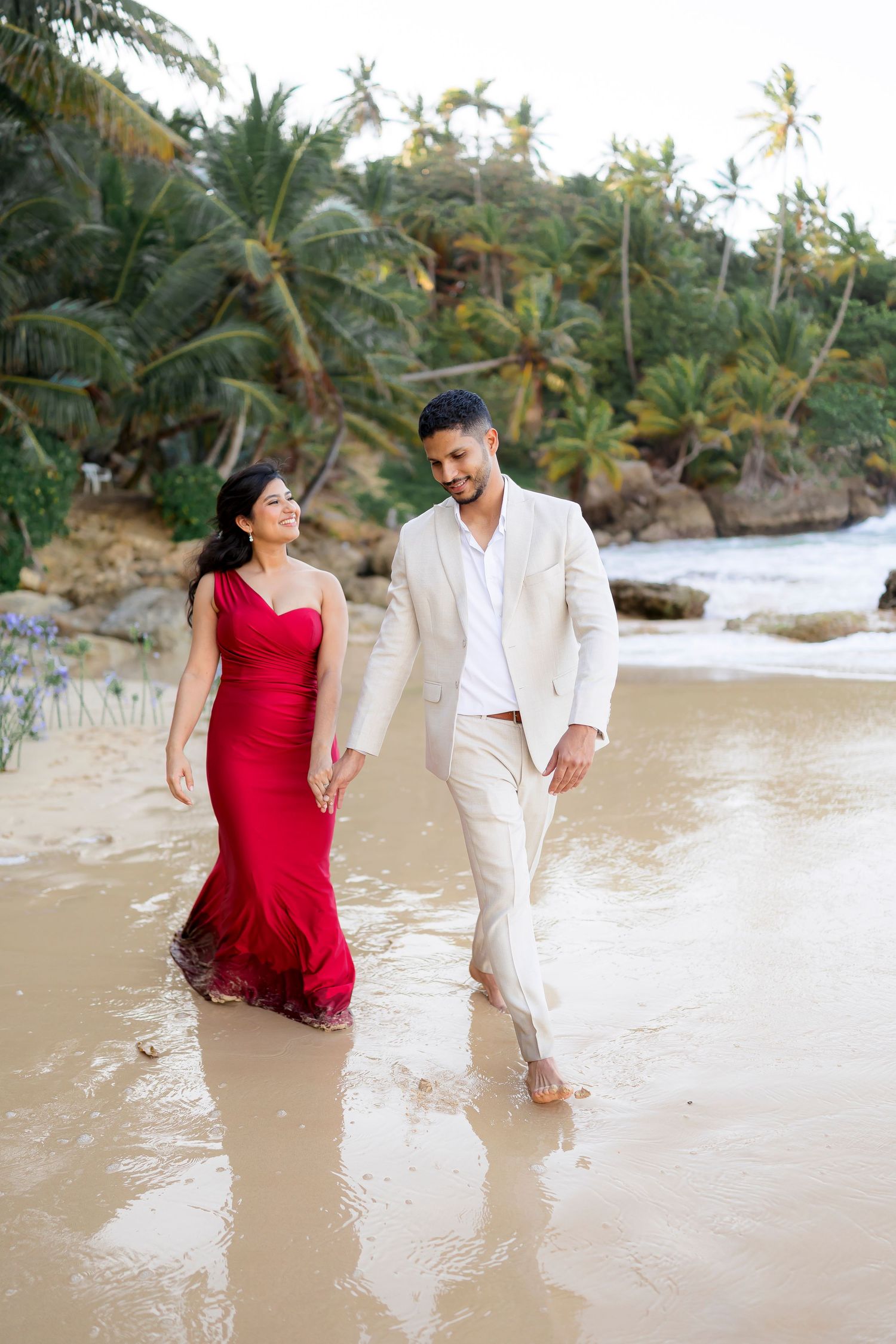 A couple walks together on a tropical beach, with a person in a red dress and another in white attire by palm trees.