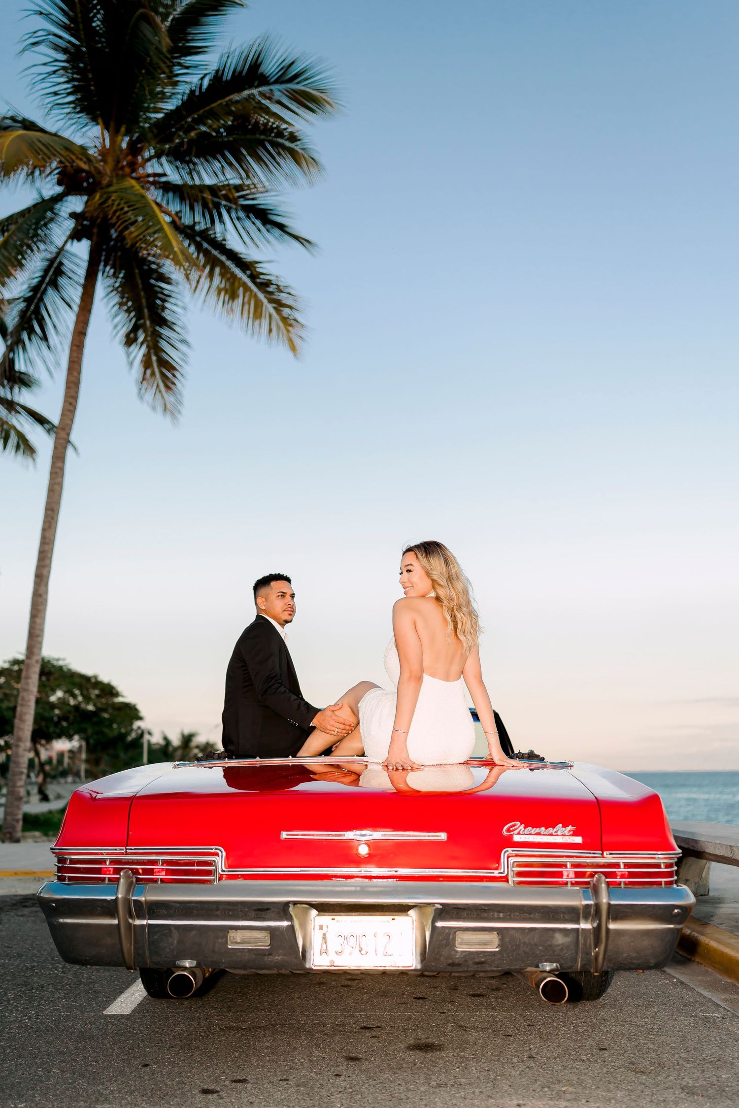 Couple sitting on red vintage car trunk with palm tree and ocean view.