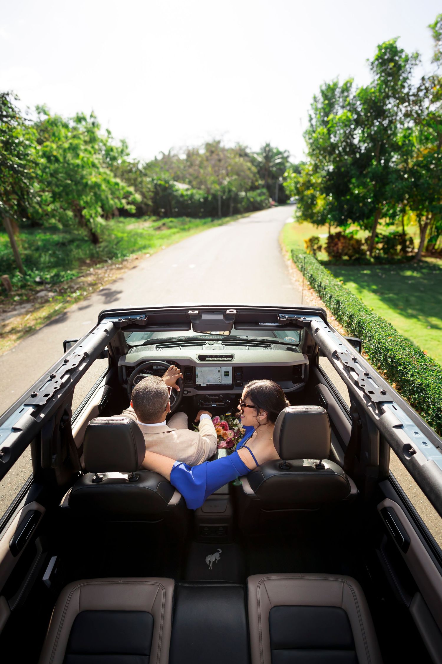 People enjoying a drive in a black convertible car on a scenic tree-lined road on a sunny day.
