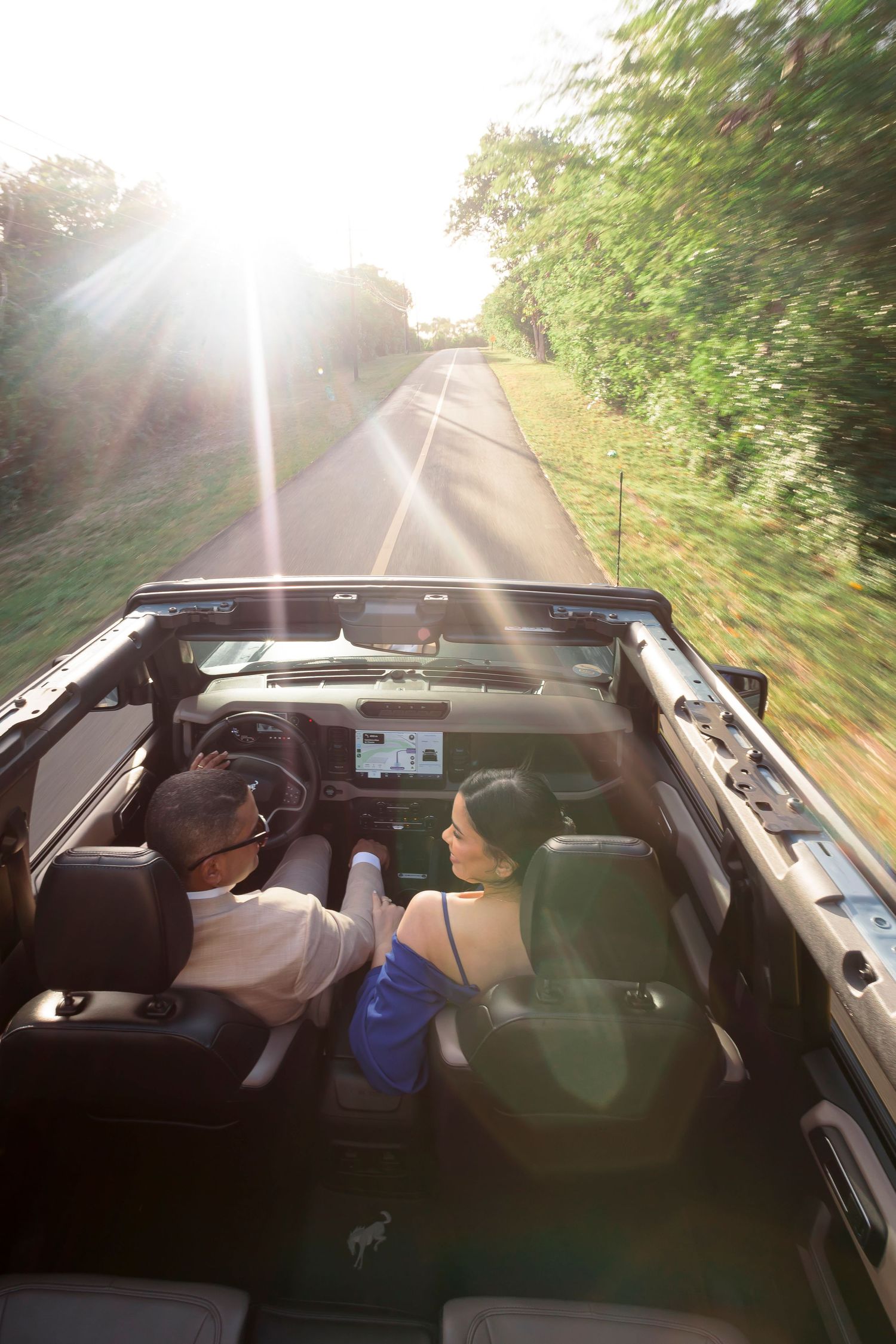 People enjoying a summer drive in a convertible car with sunlight streaming through trees along a scenic road.