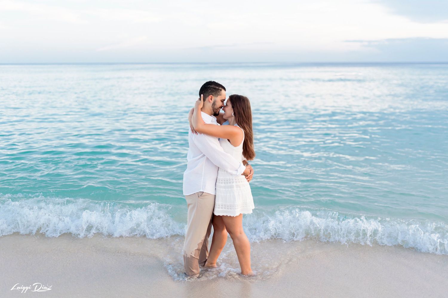 A romantic couple embraces on a beach with turquoise water in the background.