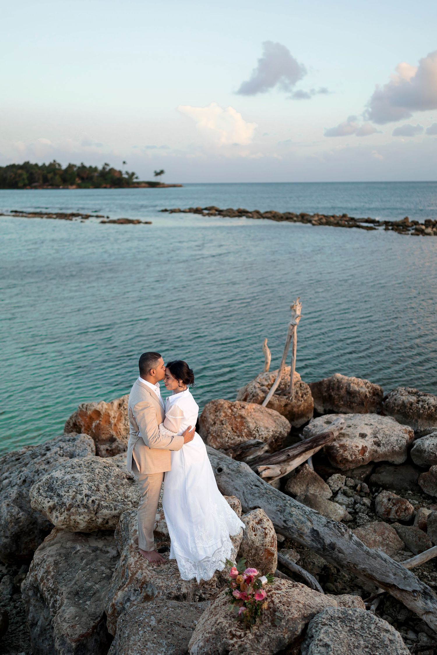 A couple embraces on rocky shoreline against turquoise ocean waters and palm tree coastline during sunset in tropical setting.