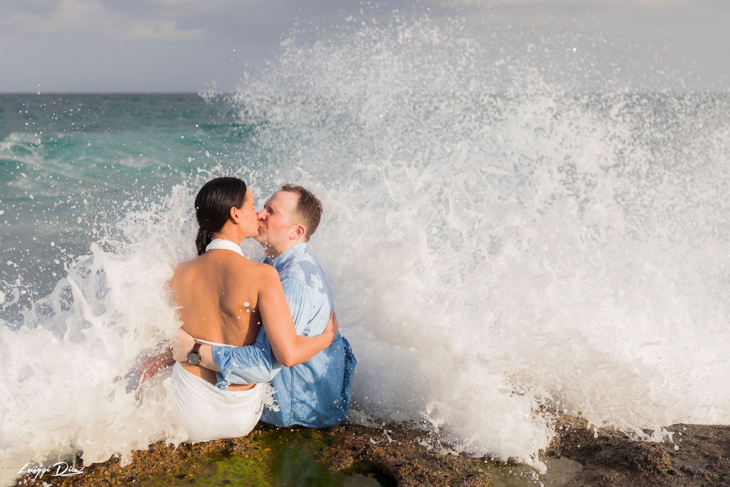 A dramatic wave crashes over a couple during their beach photoshoot.