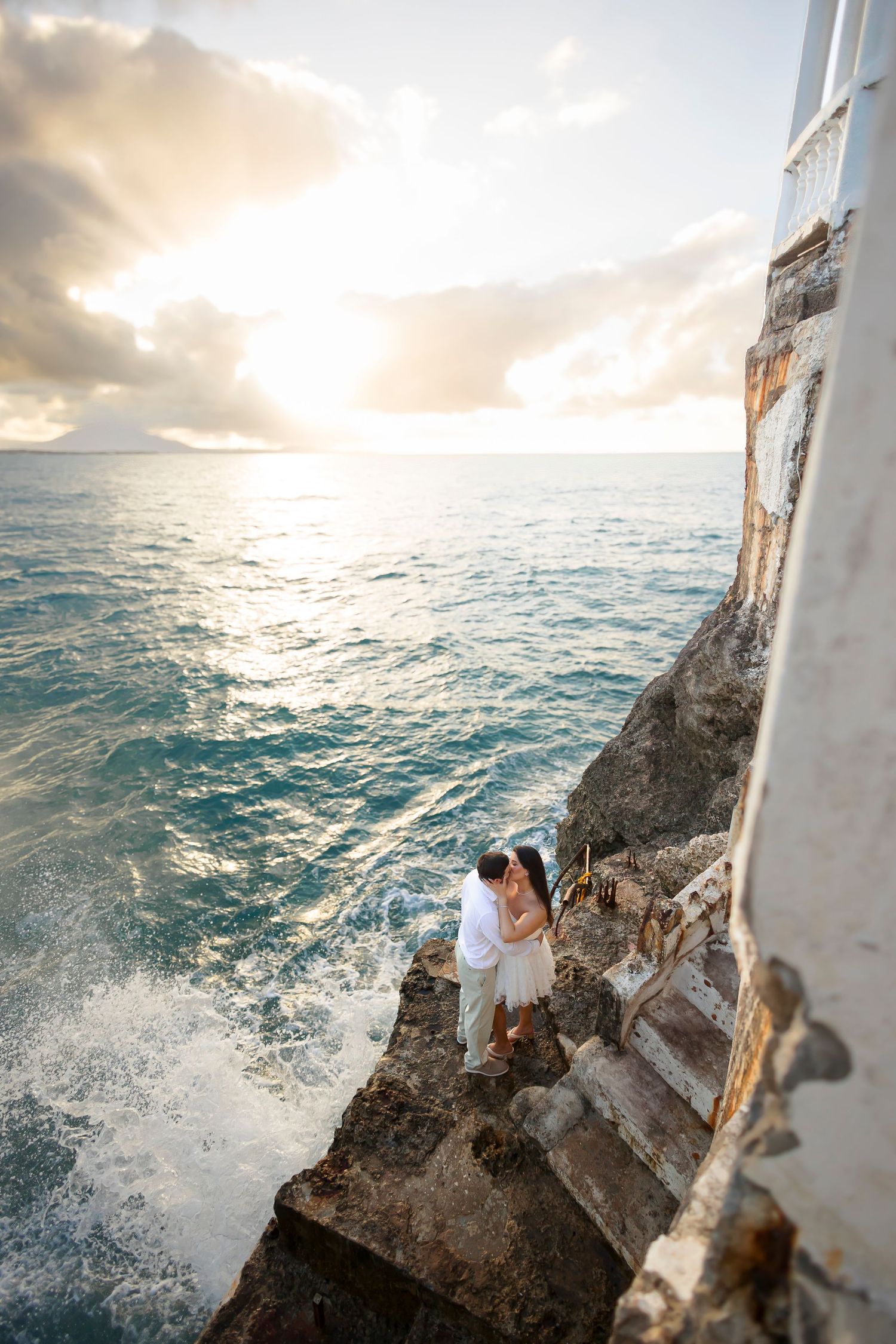 Two people embrace on a lighthouse ledge as ocean waves crash against rocks during a dramatic sunset in Hawaii.