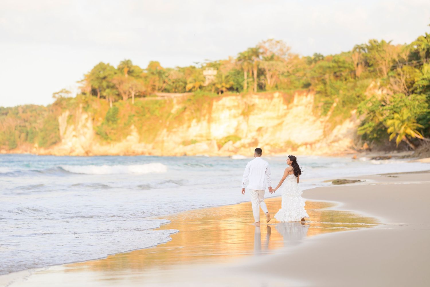 Couple walking hand in hand on sandy beach at sunset with golden cliffs in background.
