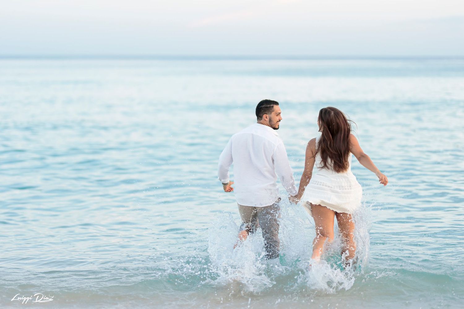 Couple splashing through ocean waves while holding hands on a beach at sunset.