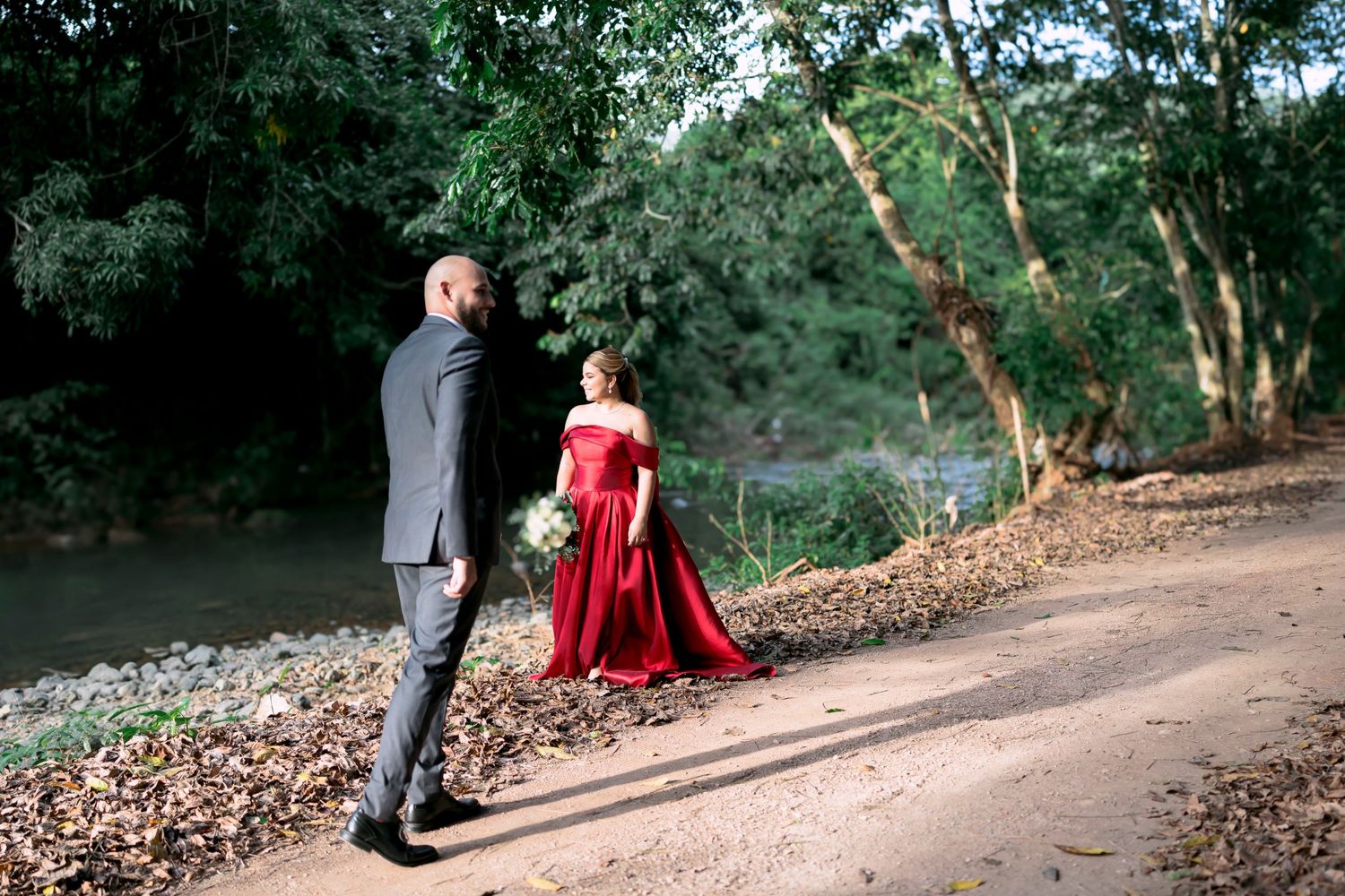 A couple walks along a wooded path, with the bride wearing a stunning red gown.