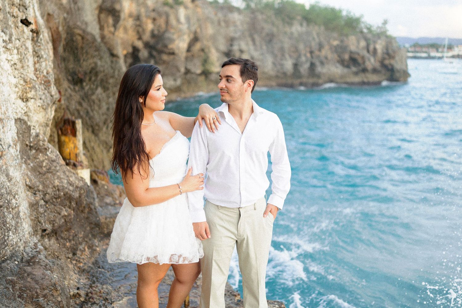 Romantic couple in white outfits standing by rocky cliffs overlooking turquoise ocean waters on a sunny day.