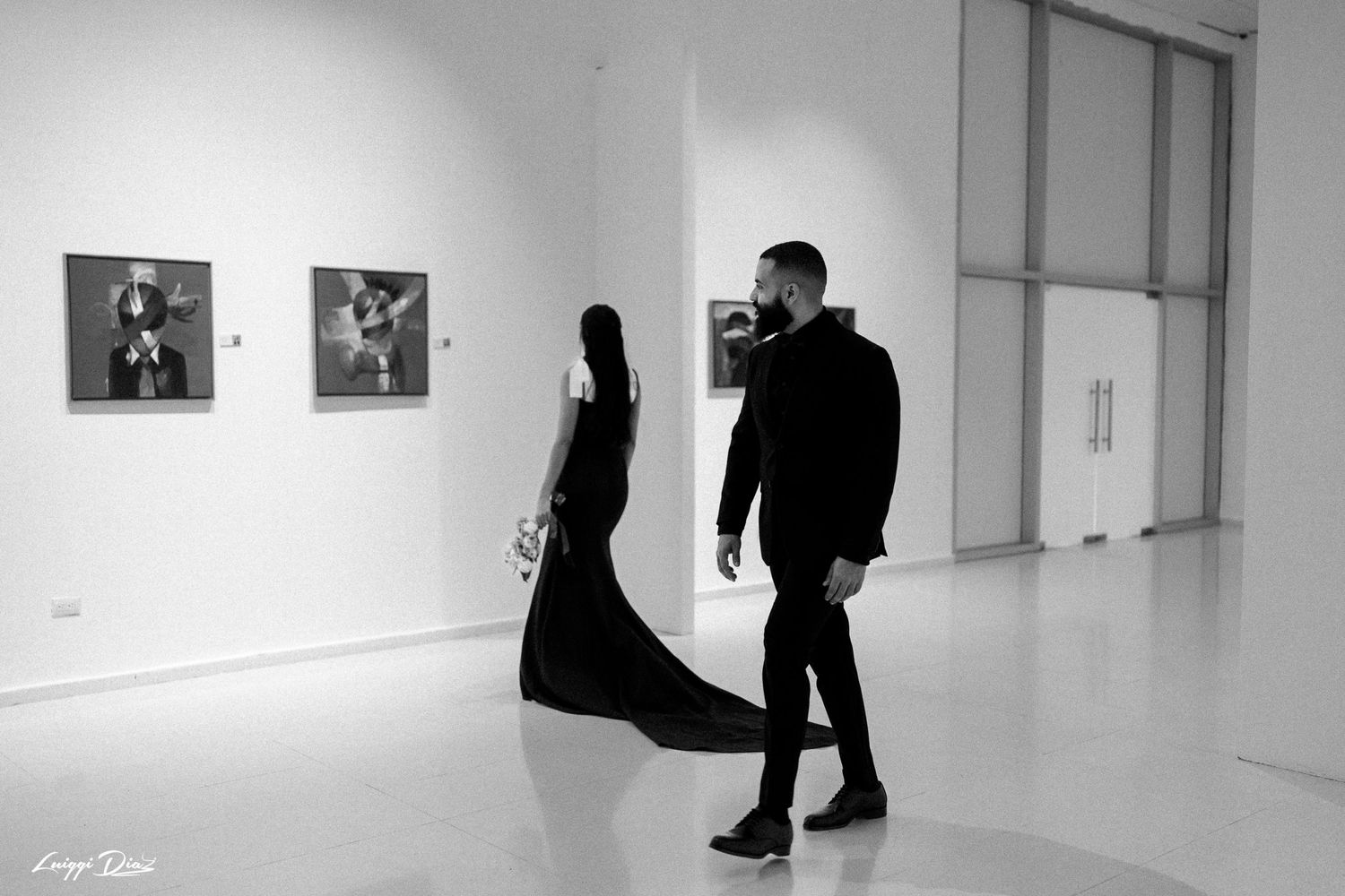 Two figures in black attire viewing art in a minimalist white gallery space.