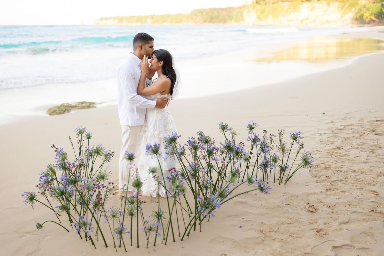 A romantic couple in white outfits embraces on a sandy beach with purple wildflowers in the foreground at sunset.
