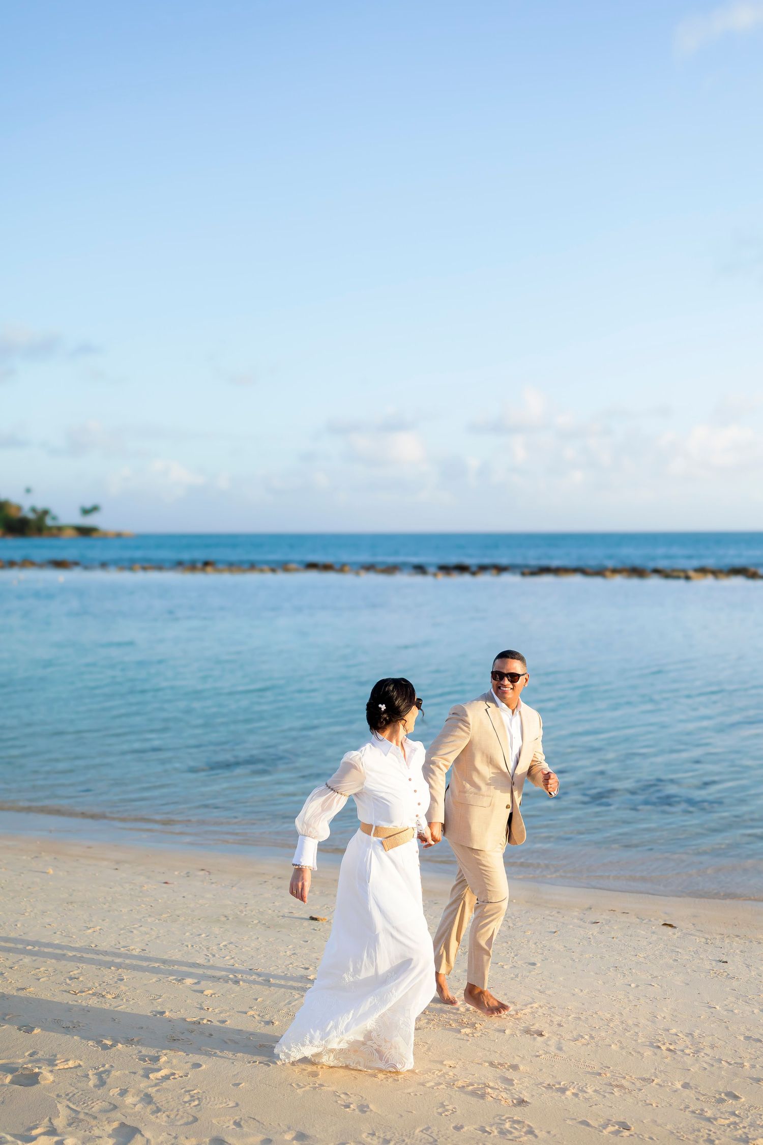 Two people in white and beige attire walking together along a tropical beach shore with turquoise waters and blue skies.