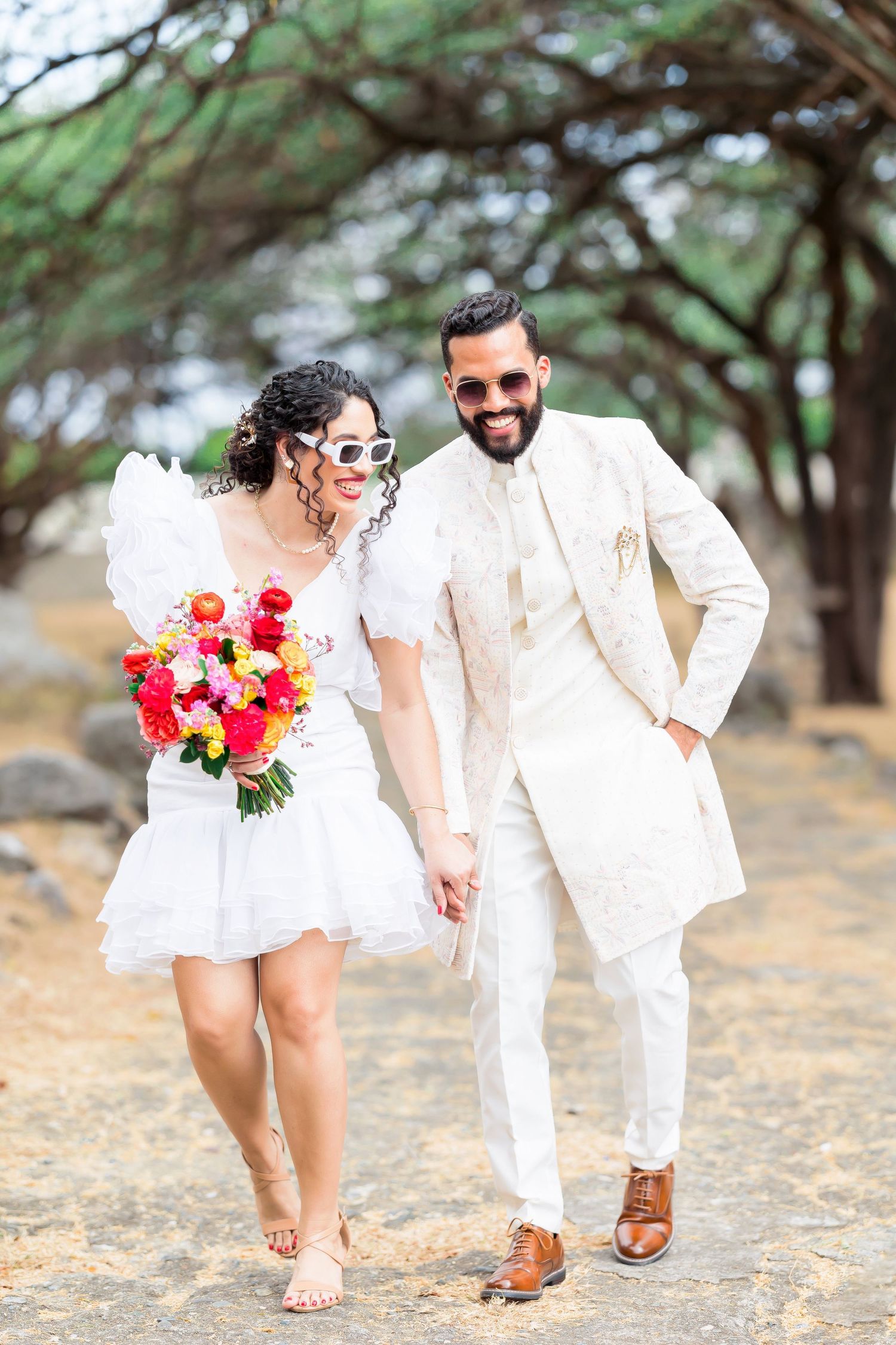 A couple in matching white outfits walk together holding a vibrant bouquet among trees.