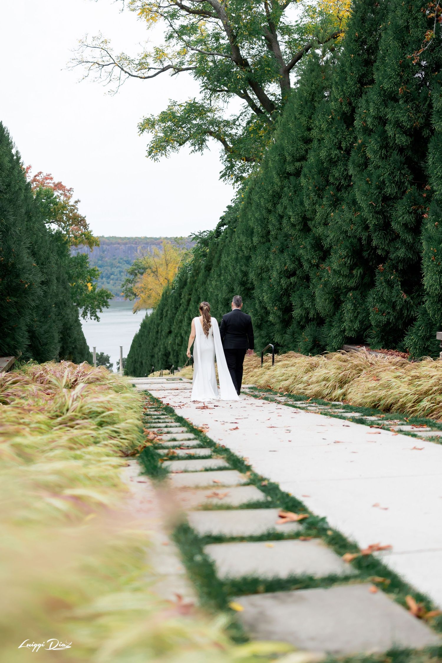 A couple walks down a stone pathway lined with tall evergreen trees near a lake.