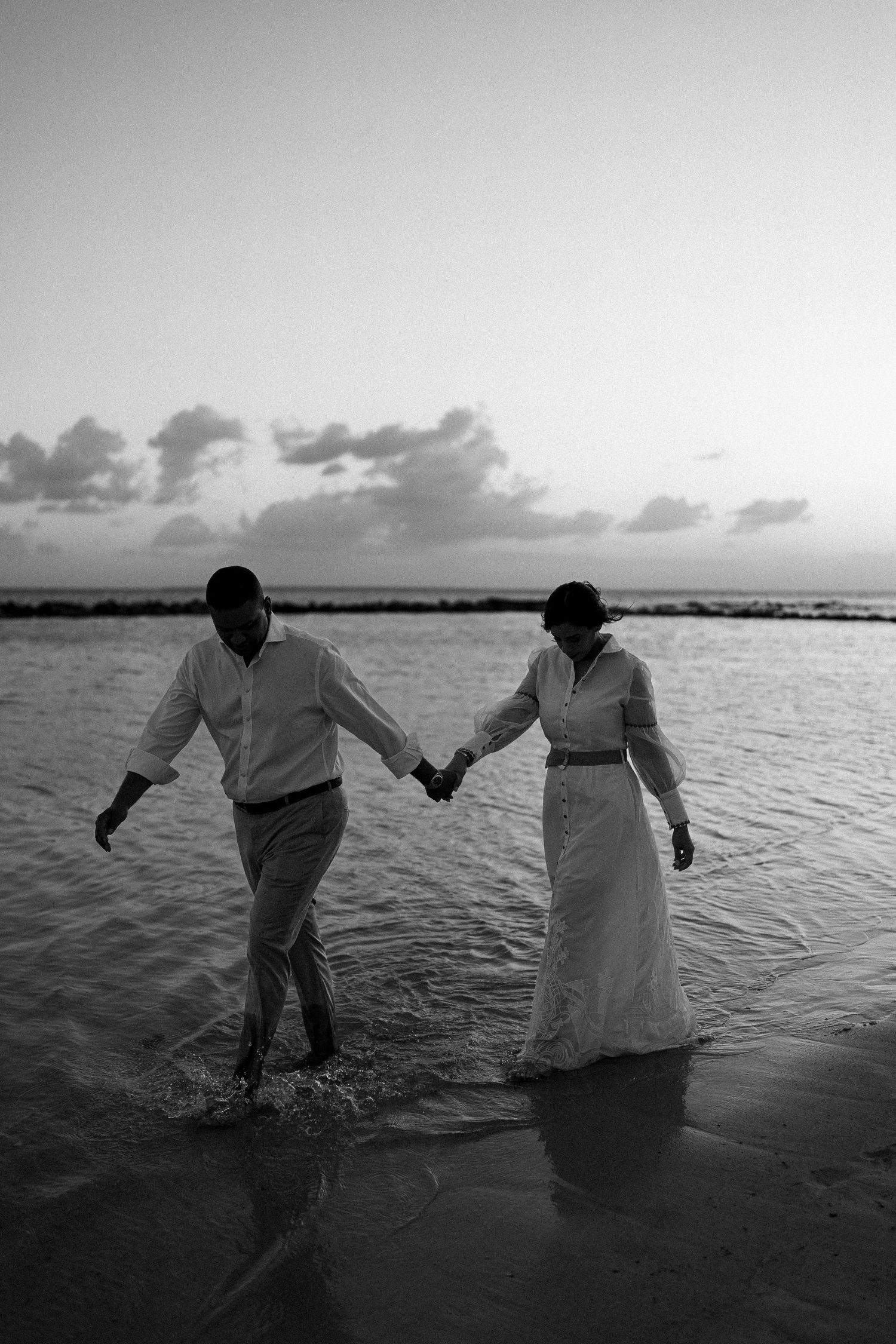A romantic black and white silhouette of a couple walking hand in hand through shallow ocean water at sunset.