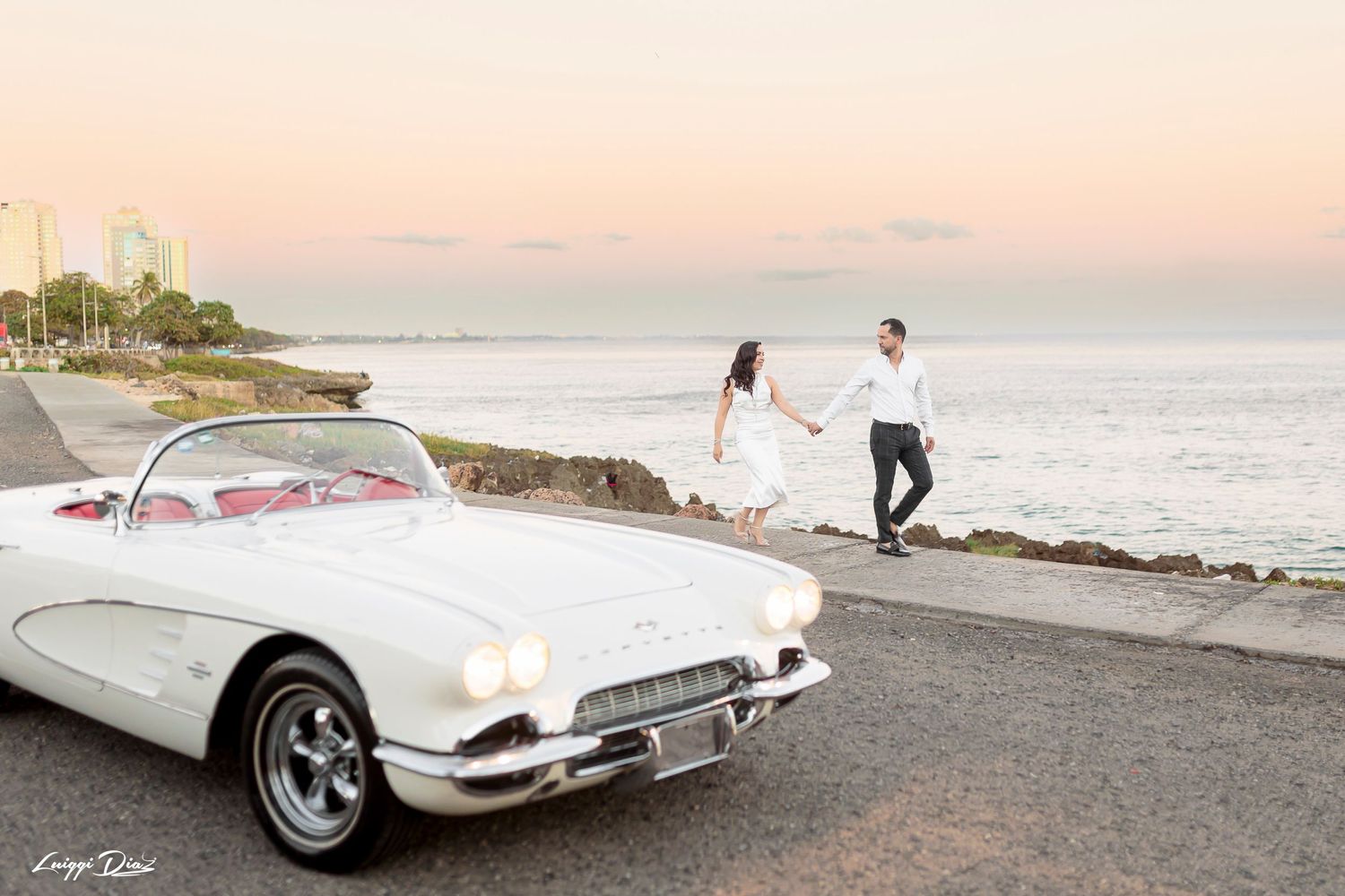 Vintage white Corvette parked near ocean with couple walking on rocky shoreline at sunset.