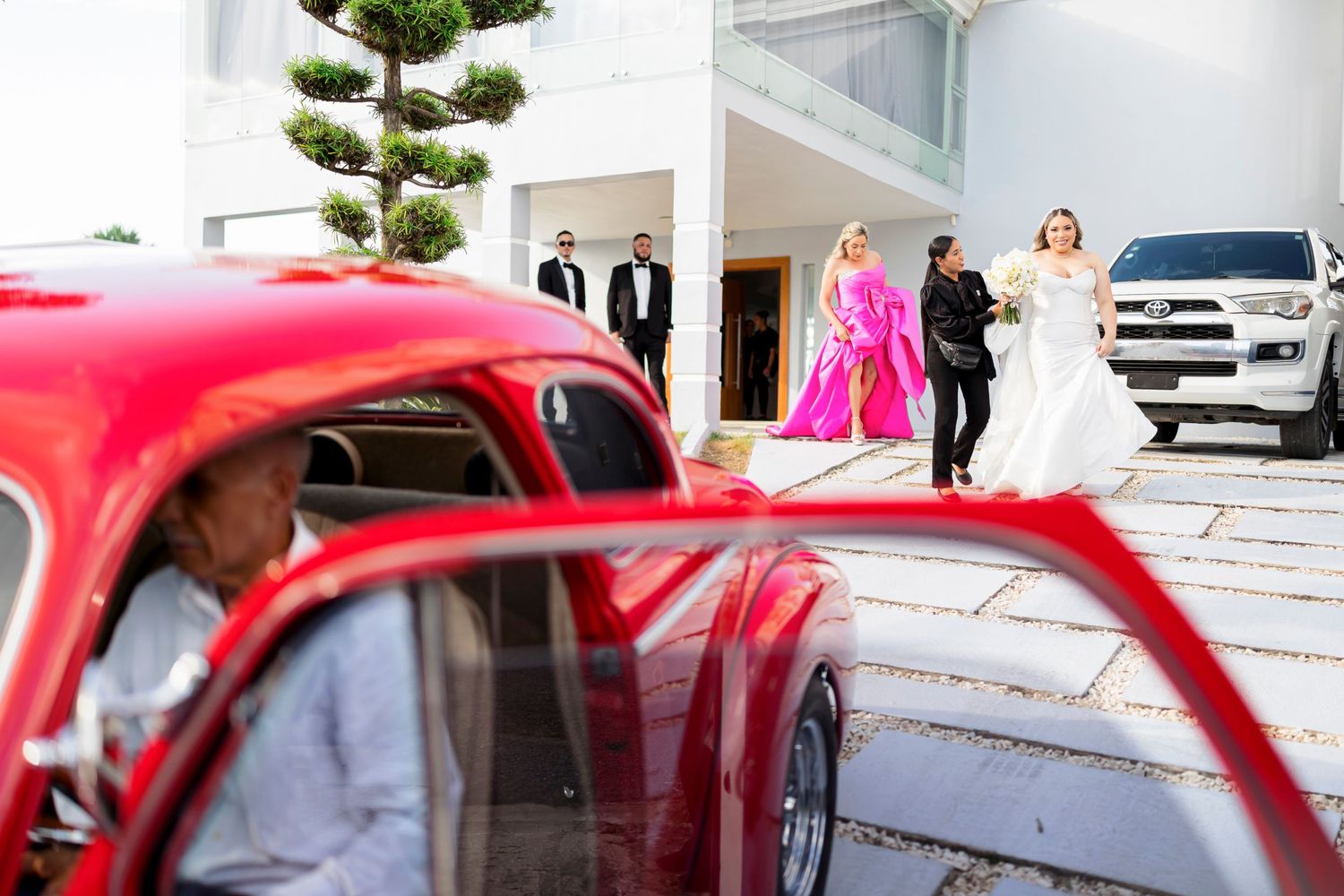 Red vintage car frames wedding party scene outside modern building.
