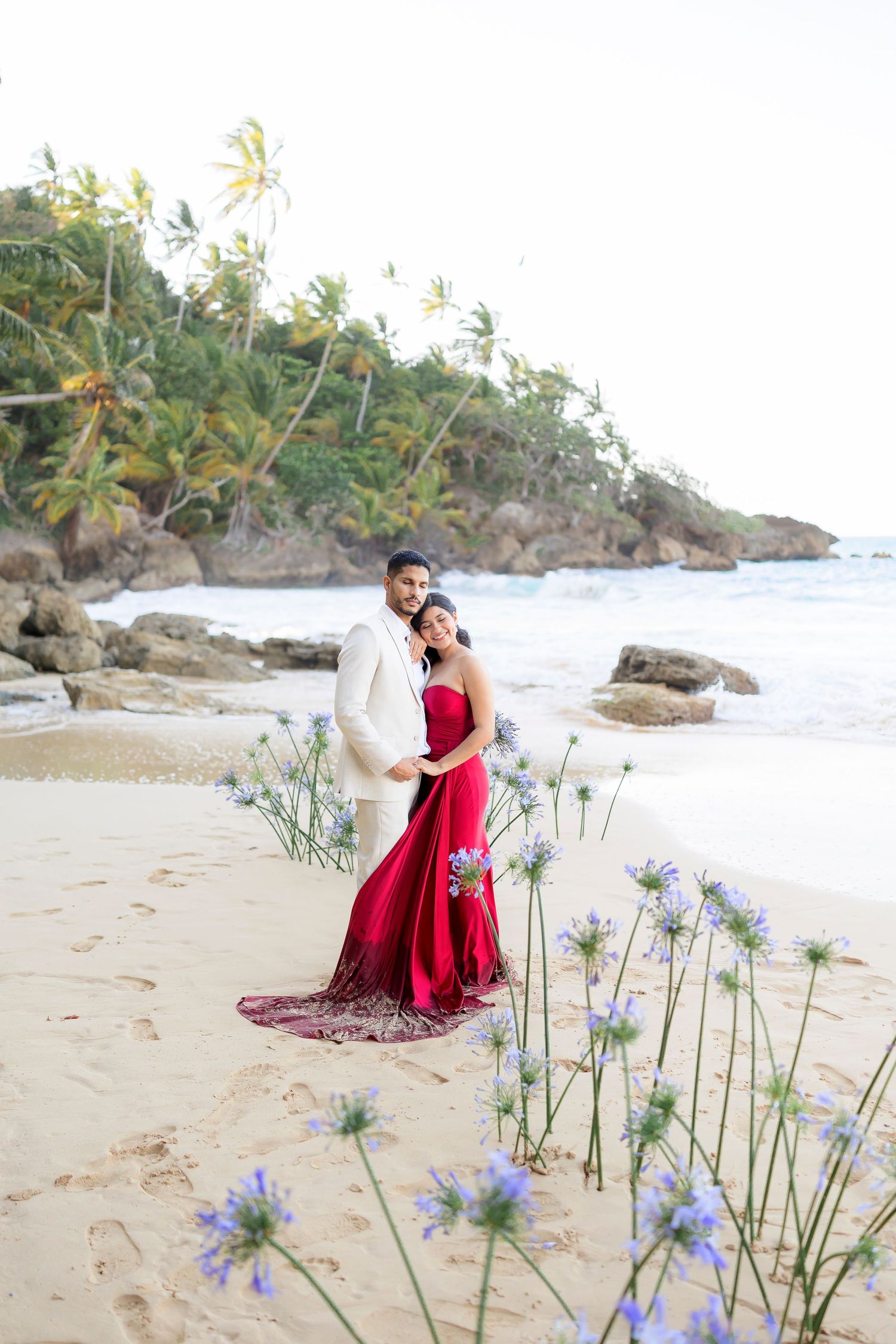 A romantic beach photoshoot with blue sea agapanthus flowers in the foreground on a tropical coastline with palm trees.