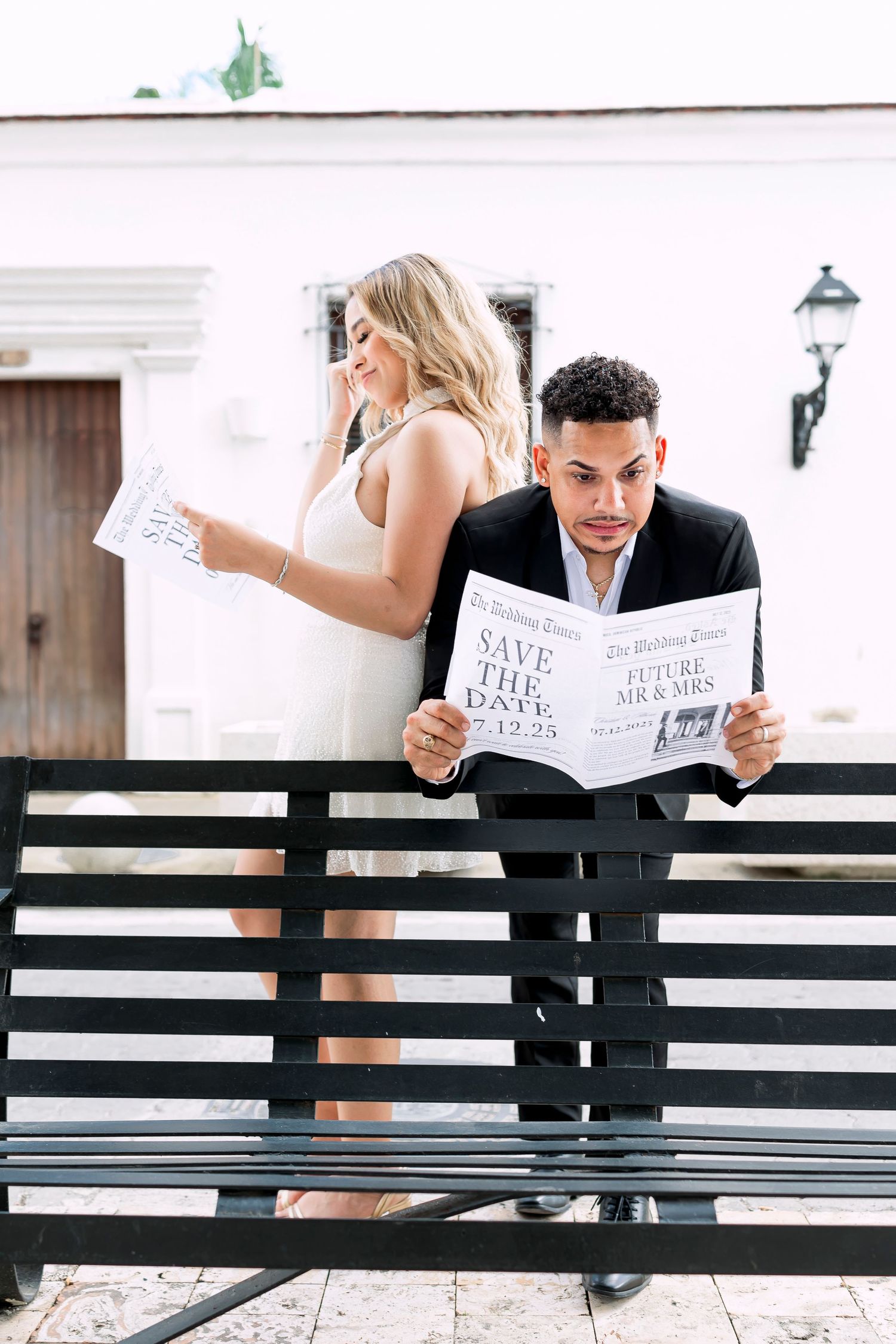 Couple reading save-the-date cards while sitting on black bench.