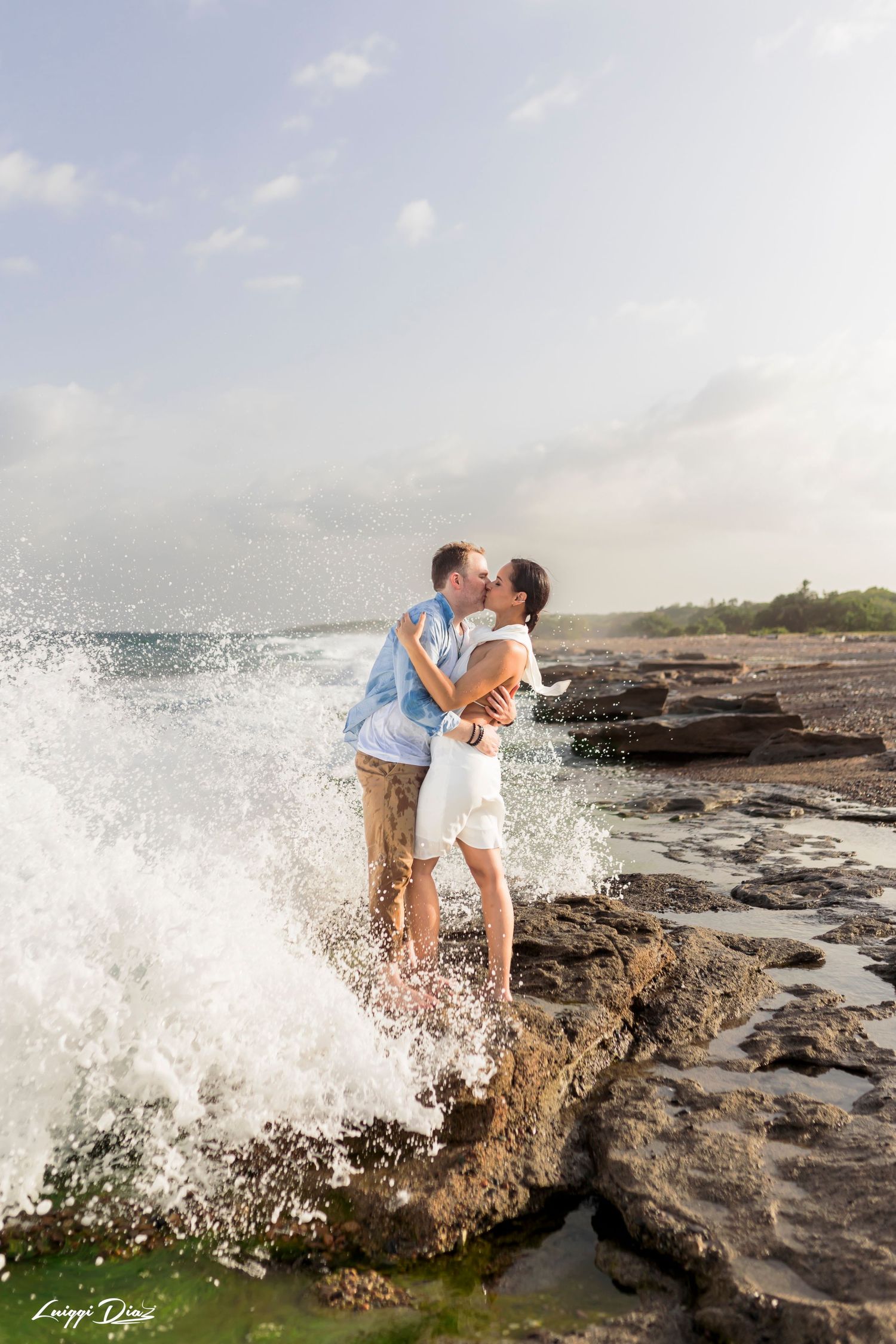 Ocean waves dramatically splash around a couple on rocky cliffs during a photoshoot.