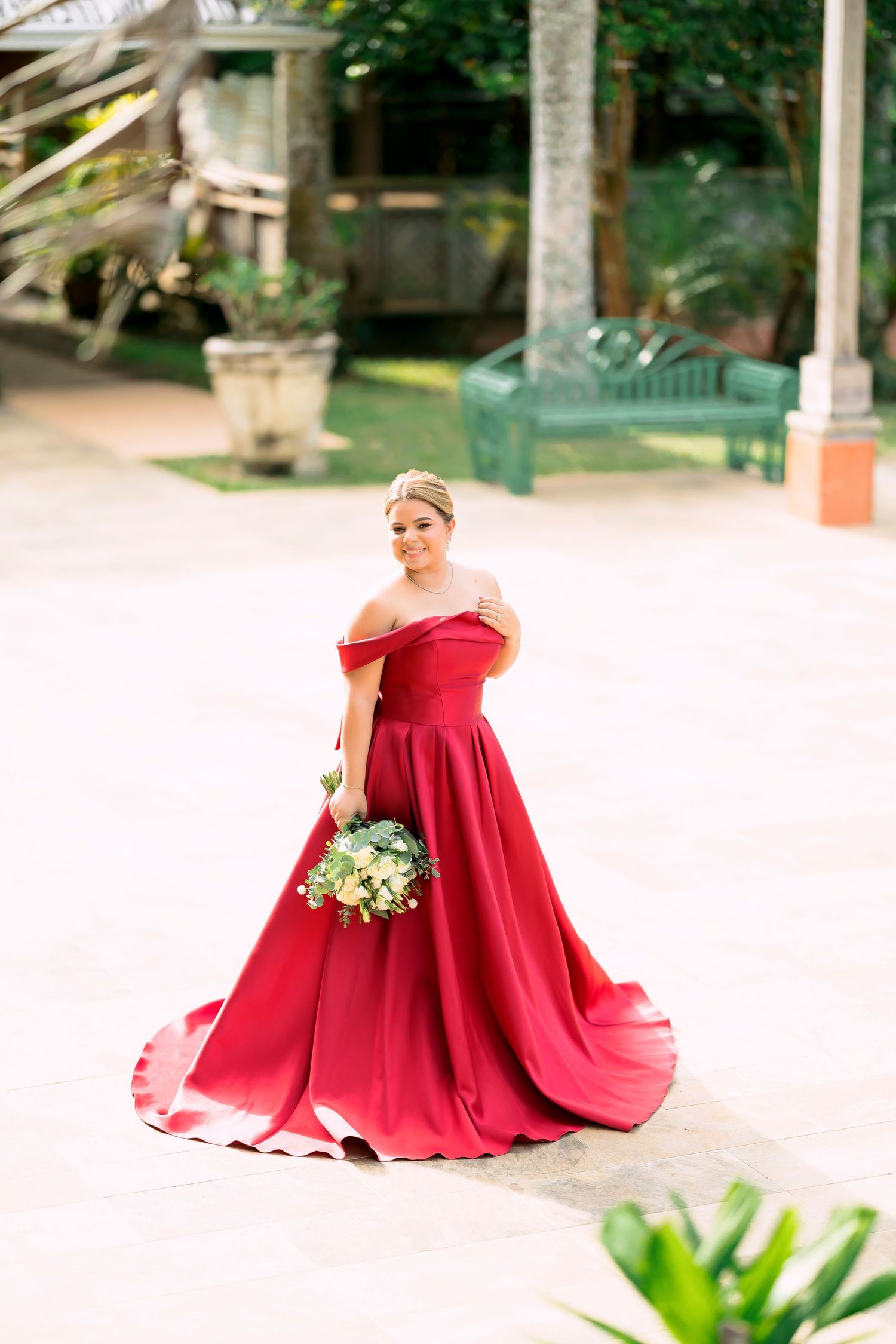 A bride in an elegant red ball gown poses in a tropical garden setting.