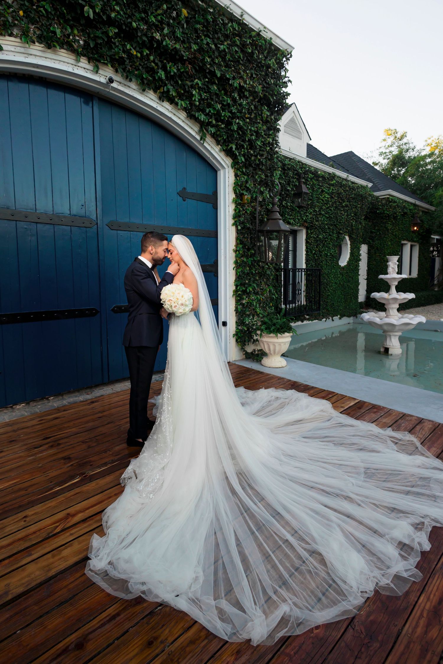 Wedding couple embracing by blue barn door with dramatic cathedral veil on wooden deck.