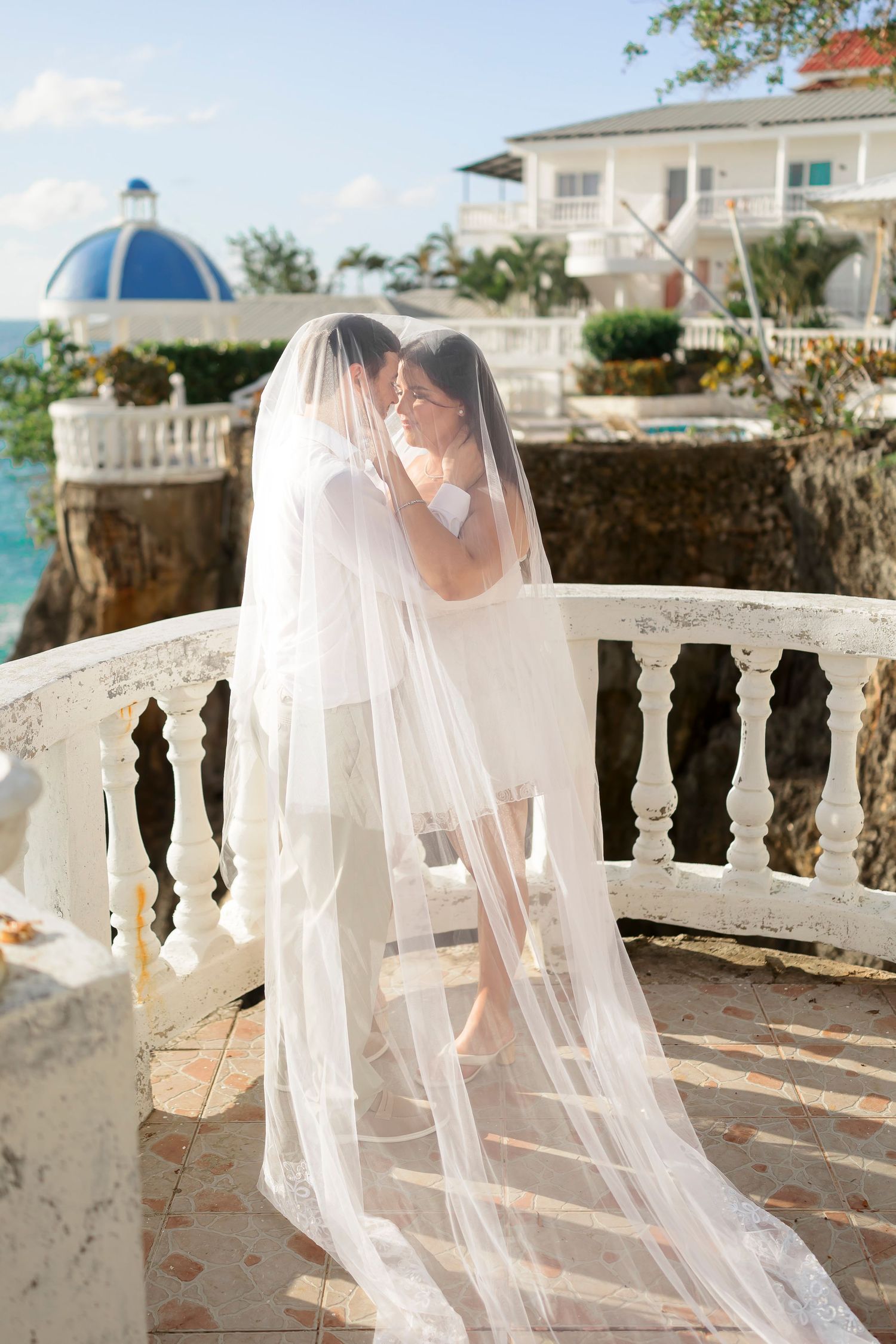 A stunning wedding photo taken at a seaside villa with ornate white stone balustrades and a domed blue structure in the background.