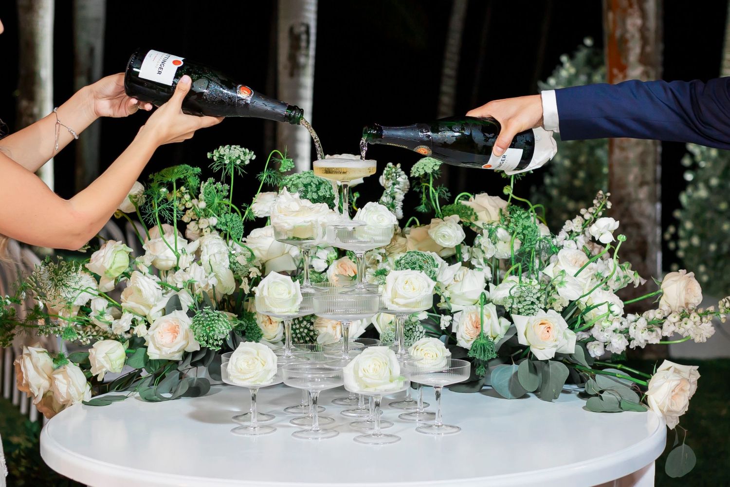 Champagne being poured over an elegant floral centerpiece with white roses and greenery.