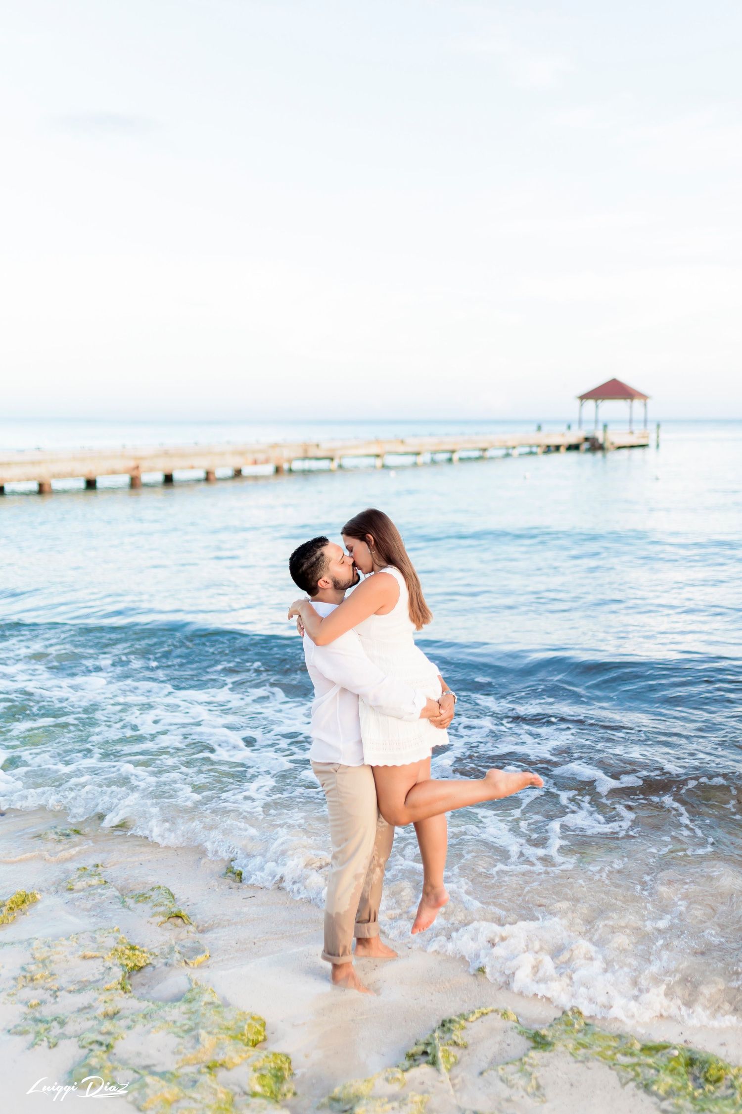 Romantic couple embracing on a tropical beach with a wooden pier and gazebo in the background.