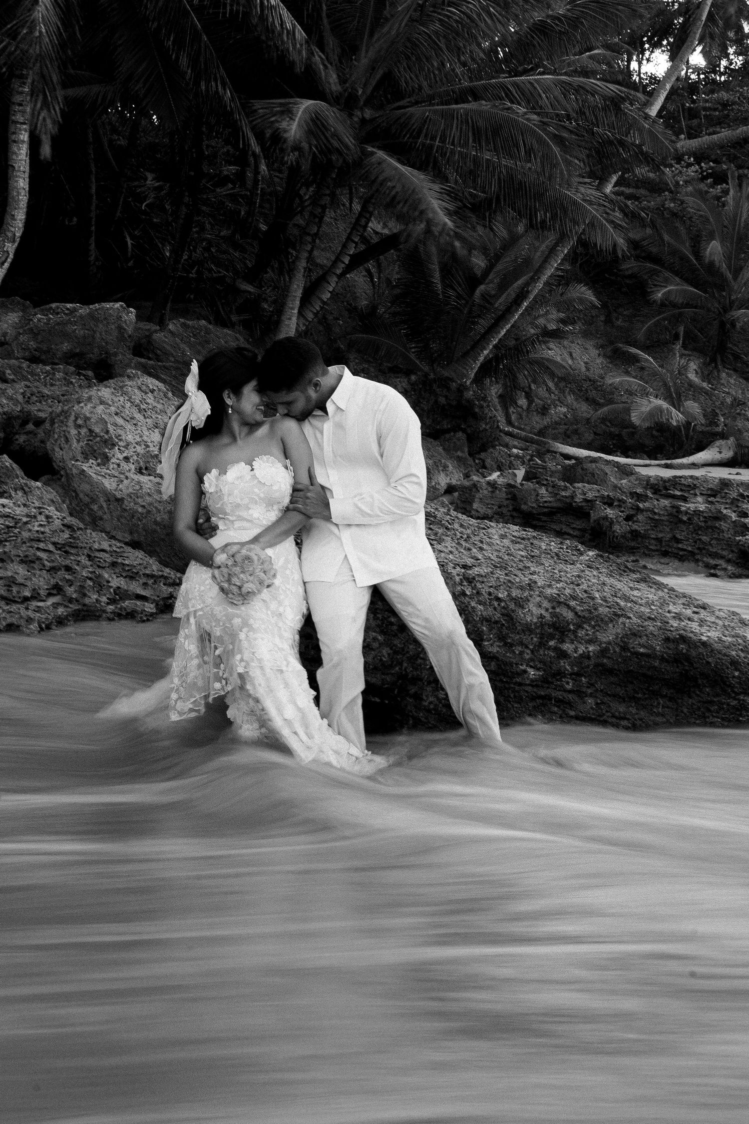 A romantic black and white wedding photo on a tropical beach with palm trees and flowing water in the foreground.