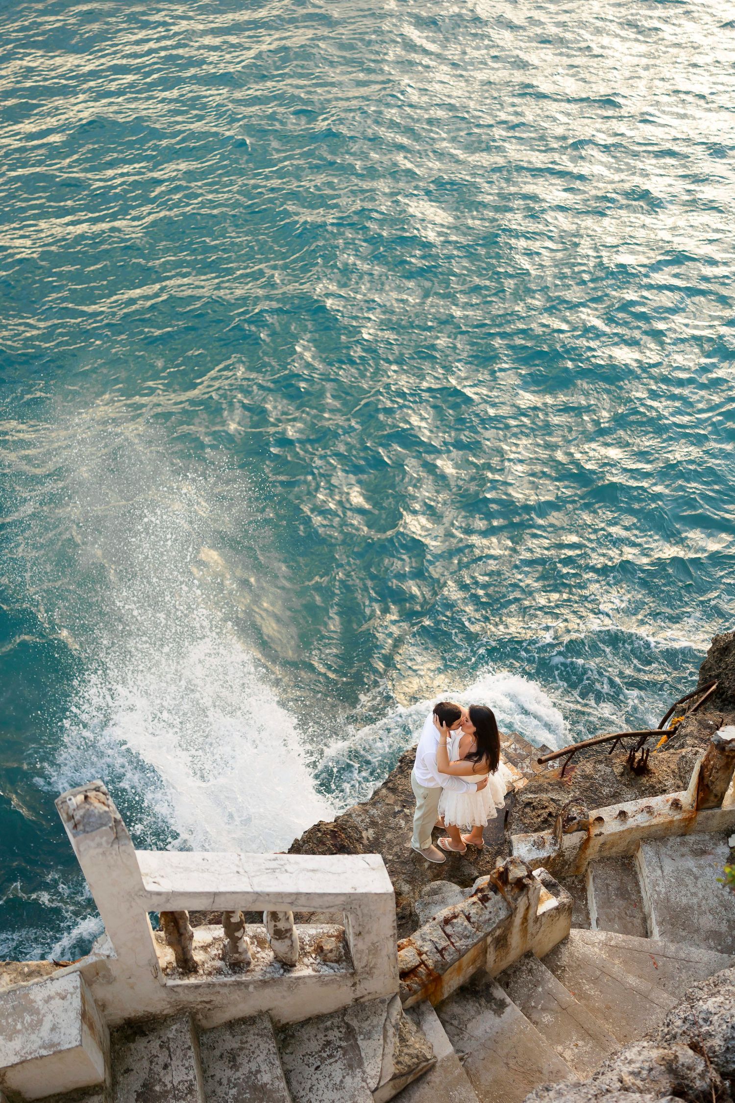 Couple embraces on stone steps overlooking dramatic ocean waves crashing against rocky cliffs at sunset.