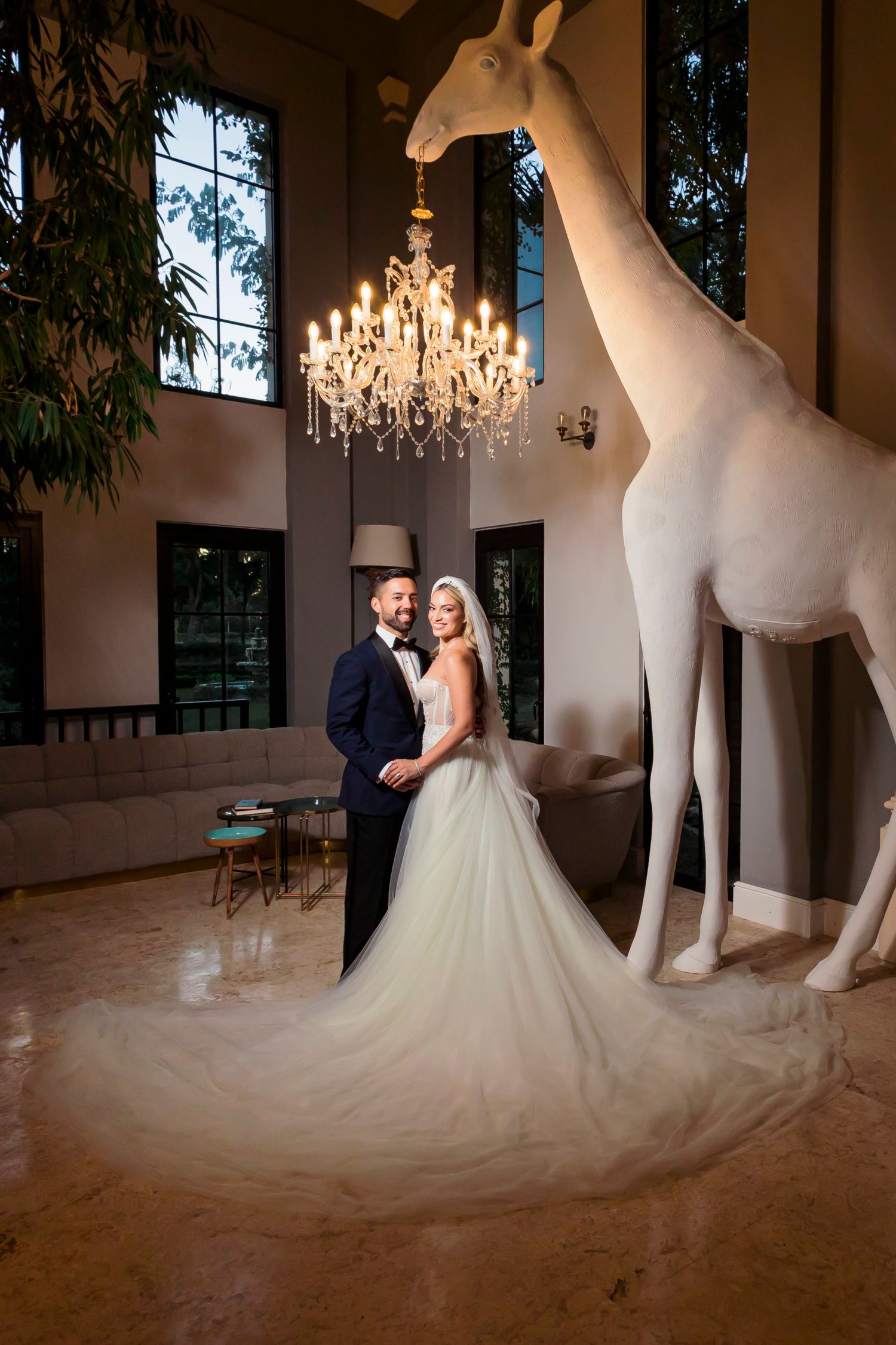 A wedding couple poses in an elegant room with a large white giraffe sculpture and crystal chandelier.
