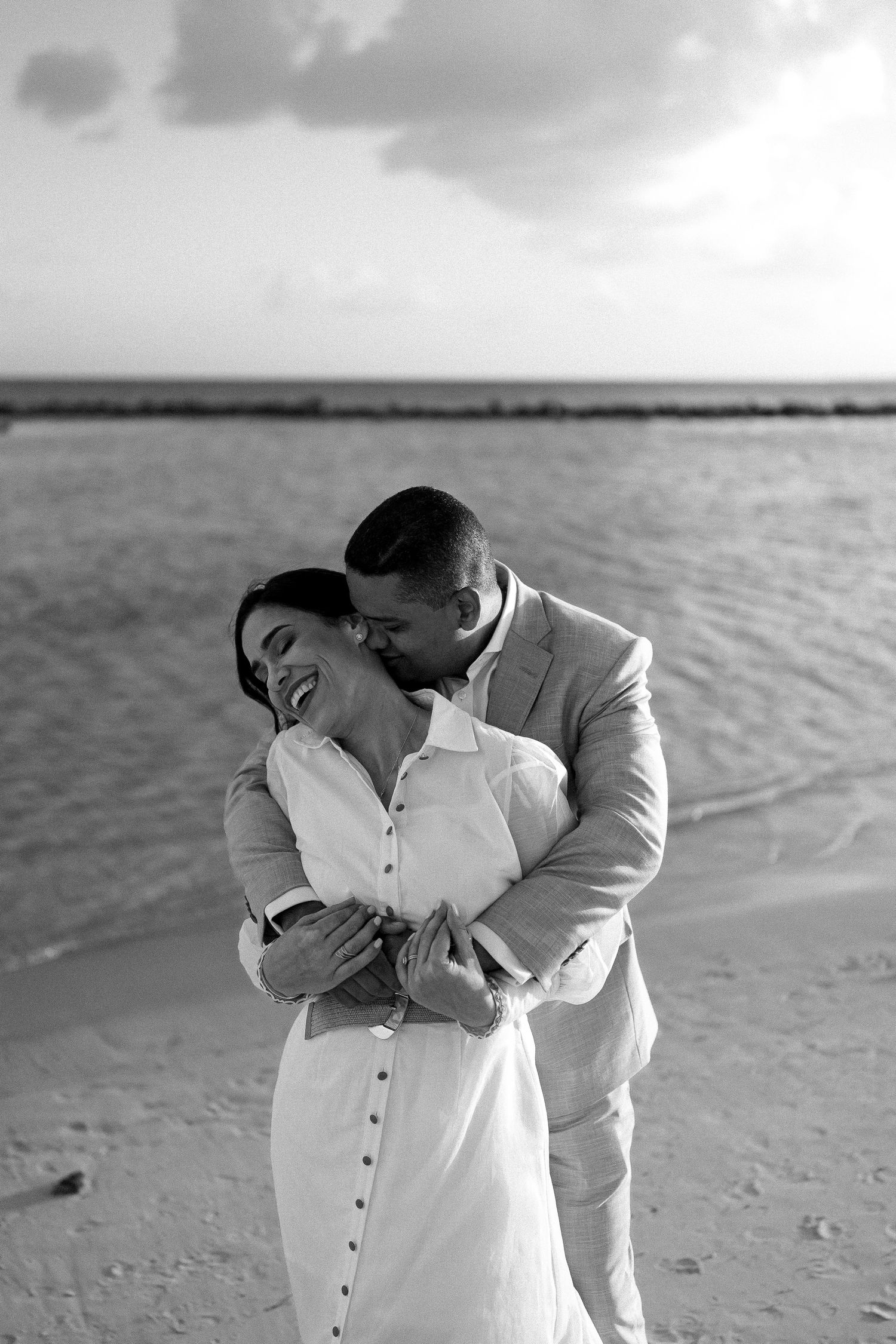 A tender black and white moment between a couple embracing on a beach with water and cloudy sky in the background.