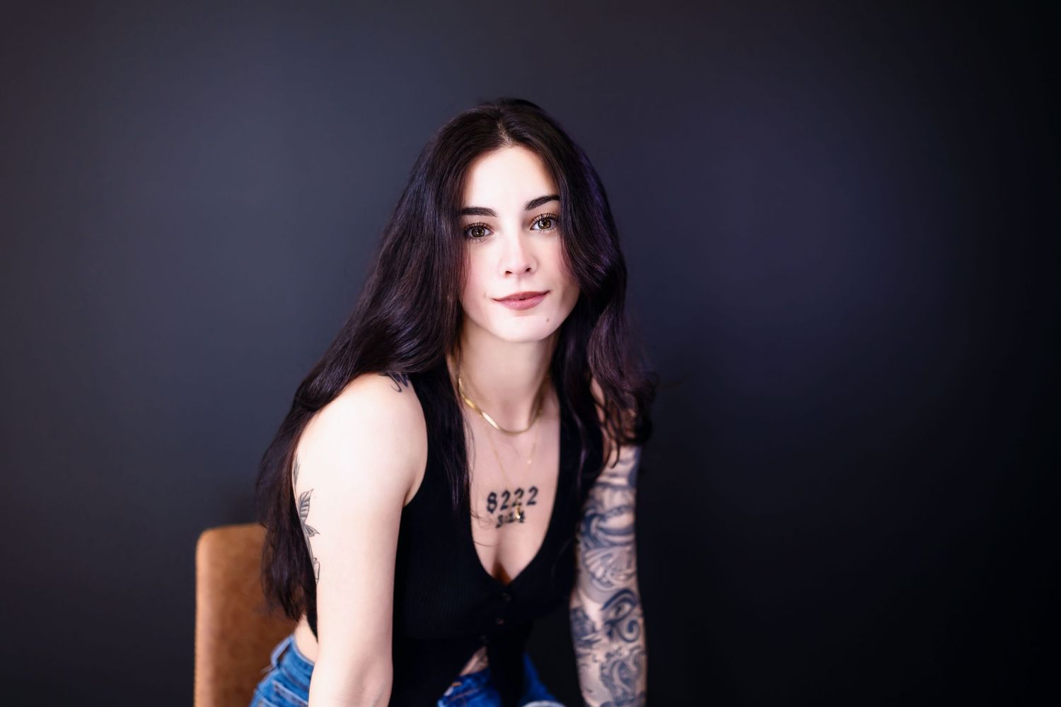Person in black tank top sits posed against dark background, showing arm tattoos and long dark hair.